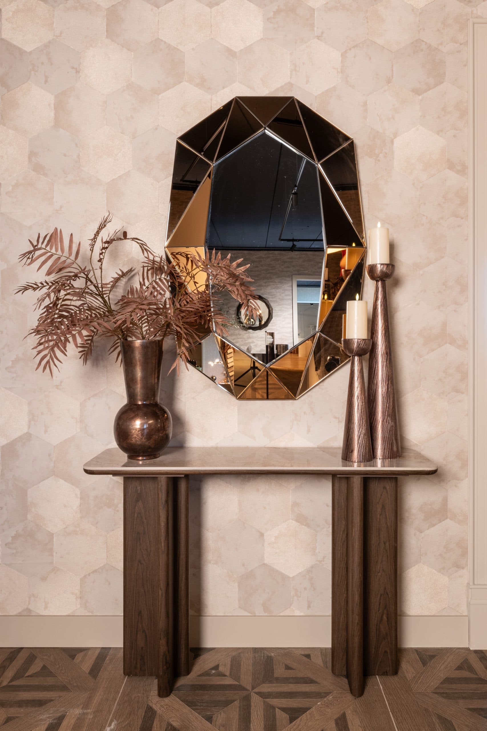 Spiegel met gefacetteerd bronskleurig frame boven een console met een bronzen vaas met gedroogde takken en twee houten kandelaars
