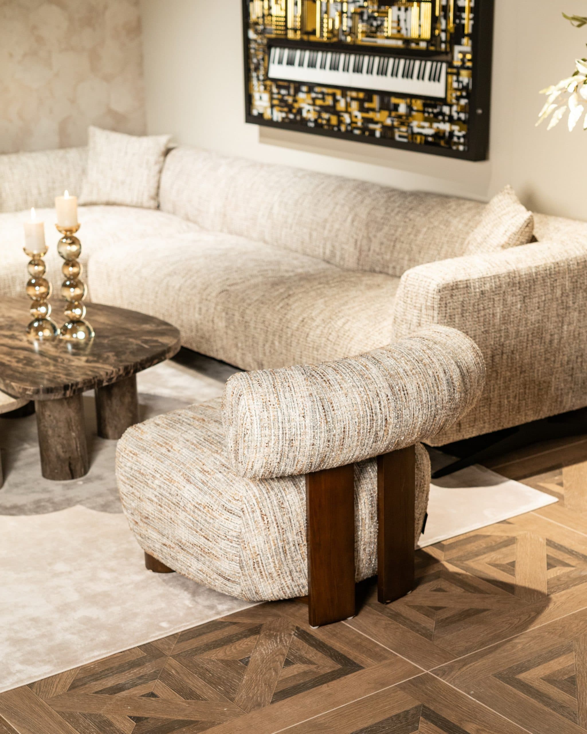 Loungestoel in gemêleerde beige tweed voor een bank in een lichtere beige stof, naast een salontafel van donker marmer met gouden kandelaars