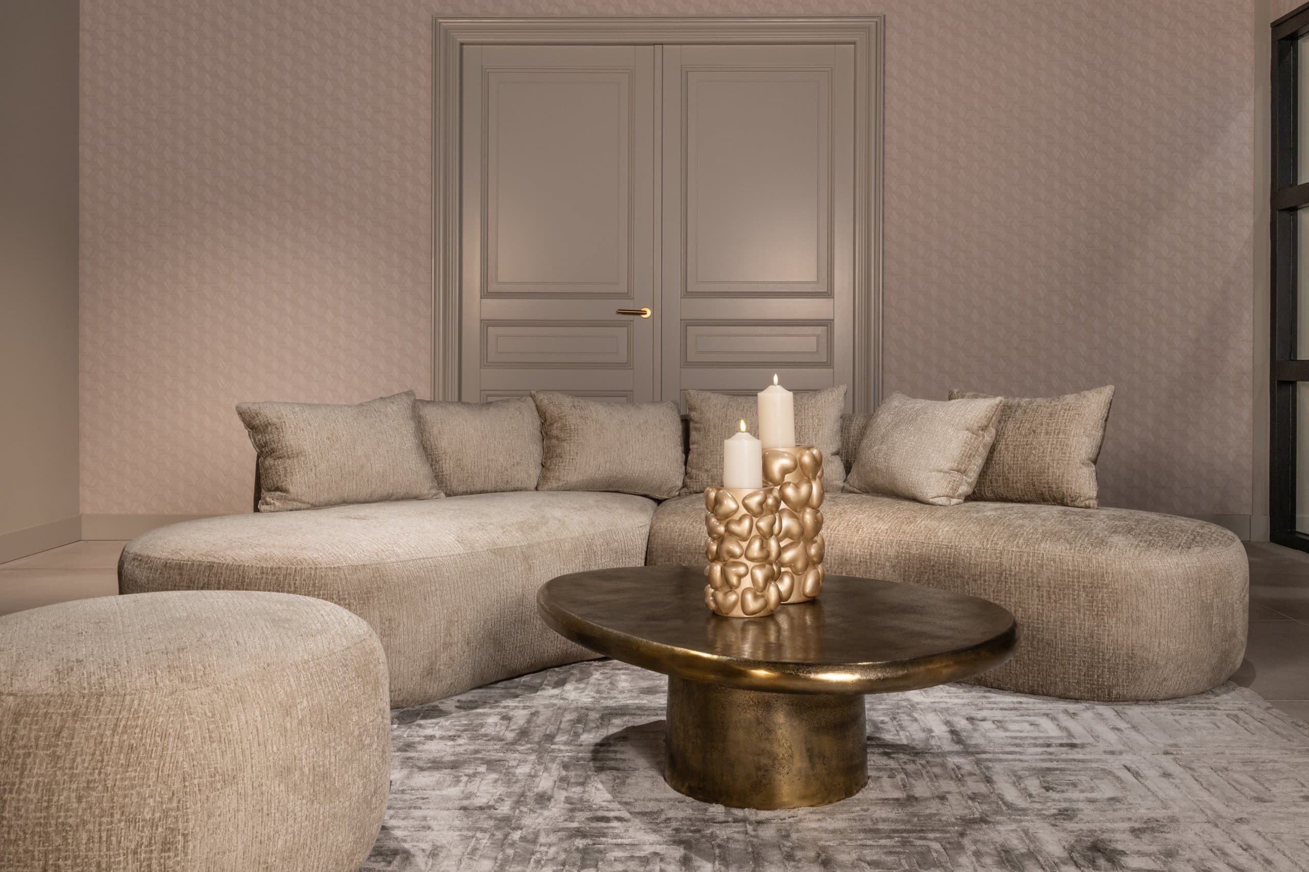 Beige hoekbank met losse kussens rond een ronde goudkleurige salontafel met twee brandende kaarsen, in een kamer met lichte paneeldeuren