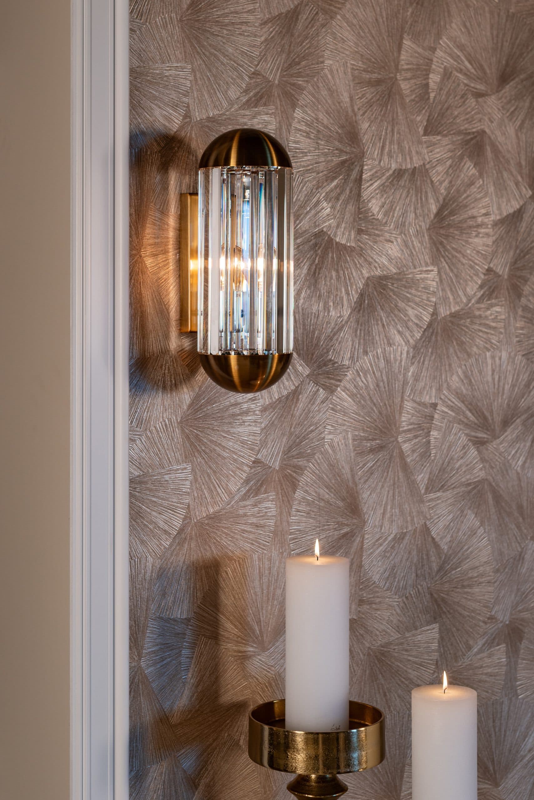 Verlichte wandlamp met glazen staafjes en gouden voet tegen een wand met grafisch bladpatroon