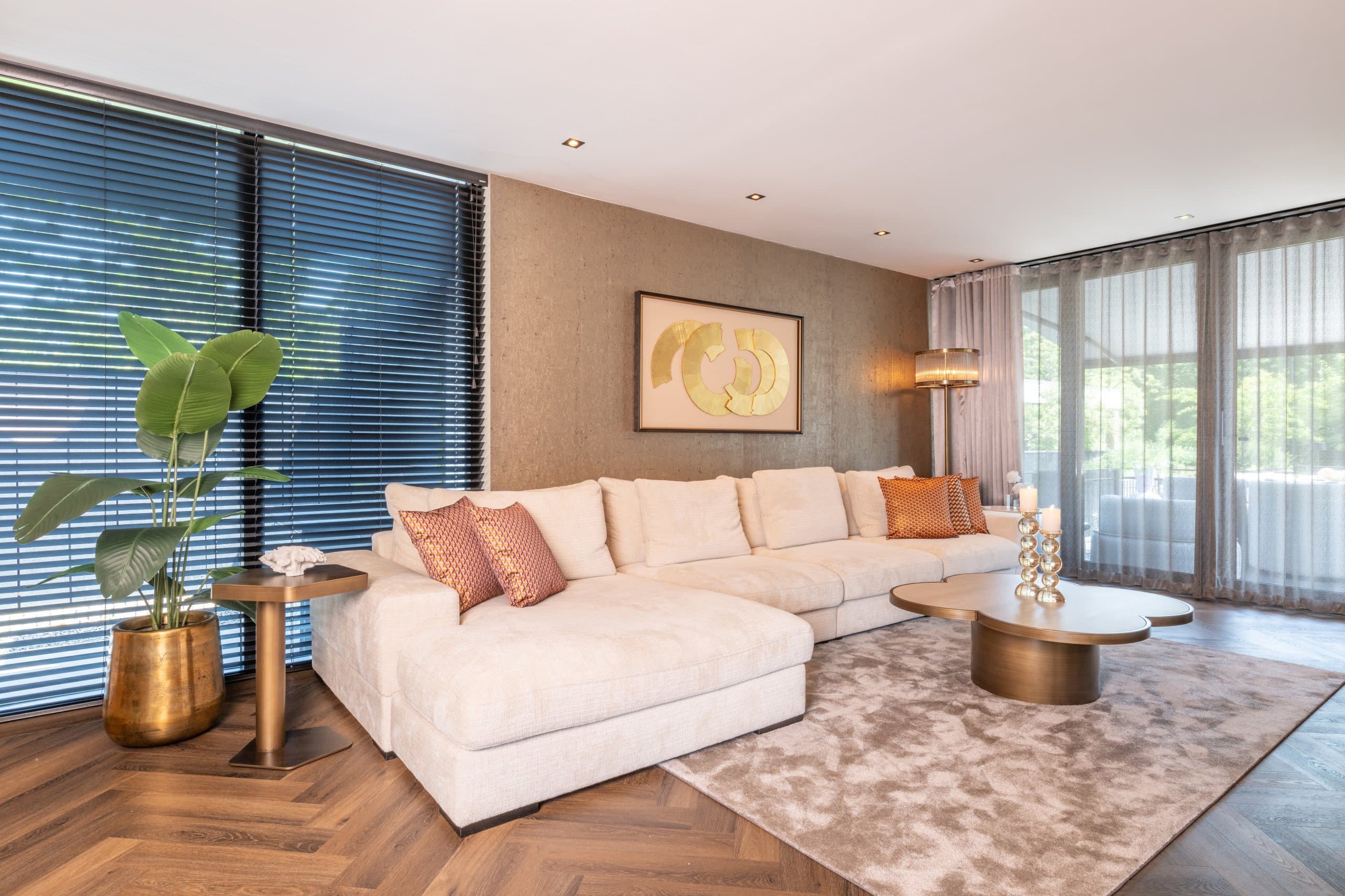 Woonkamer met crèmekleurige hoekbank, een grote groene plant in een gouden pot bij het raam en een staande lamp naast de bank