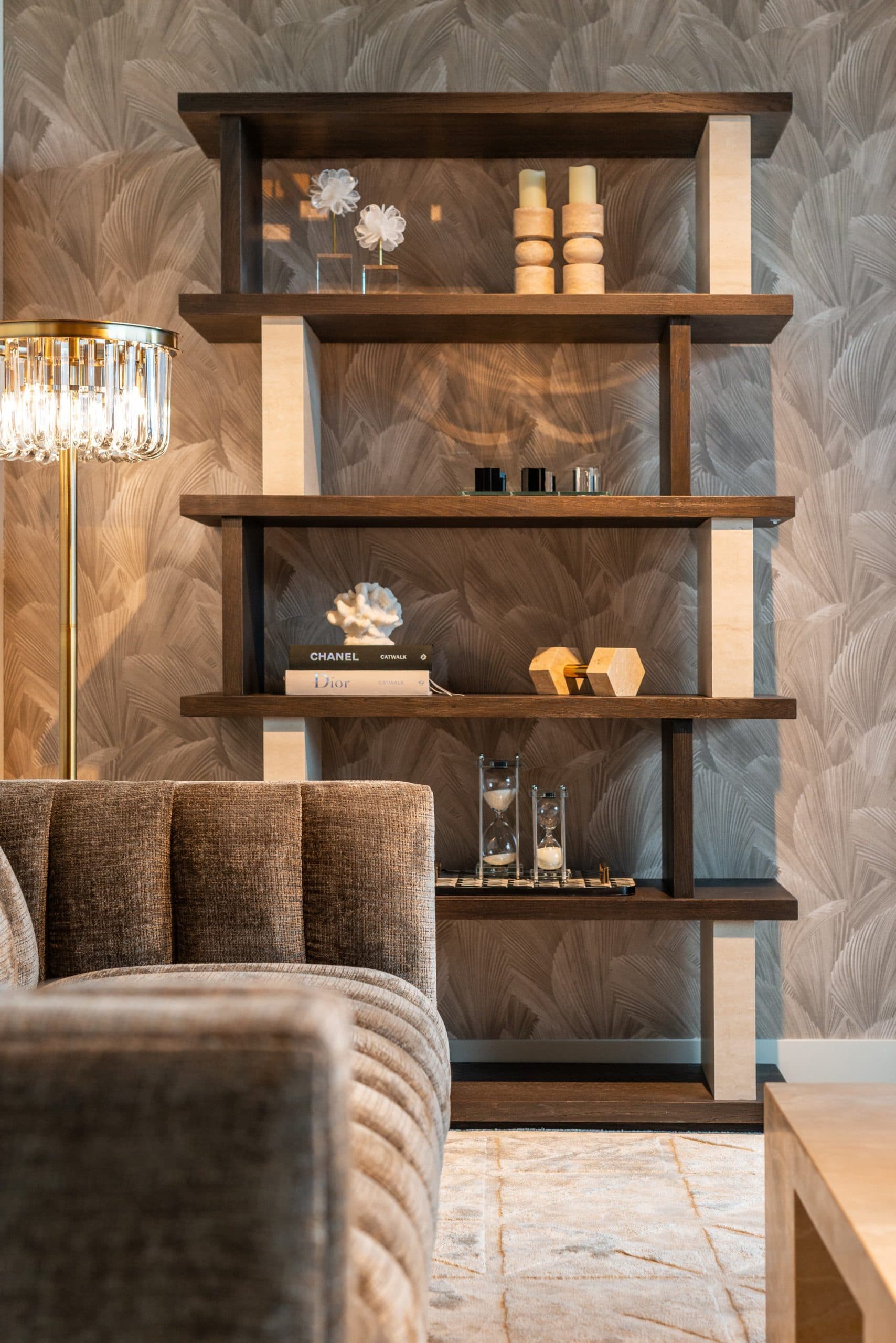 Donkerhouten wandkast met koraalobject, boeken en twee zandlopers, warm aangelicht door een gouden vloerlamp links in beeld