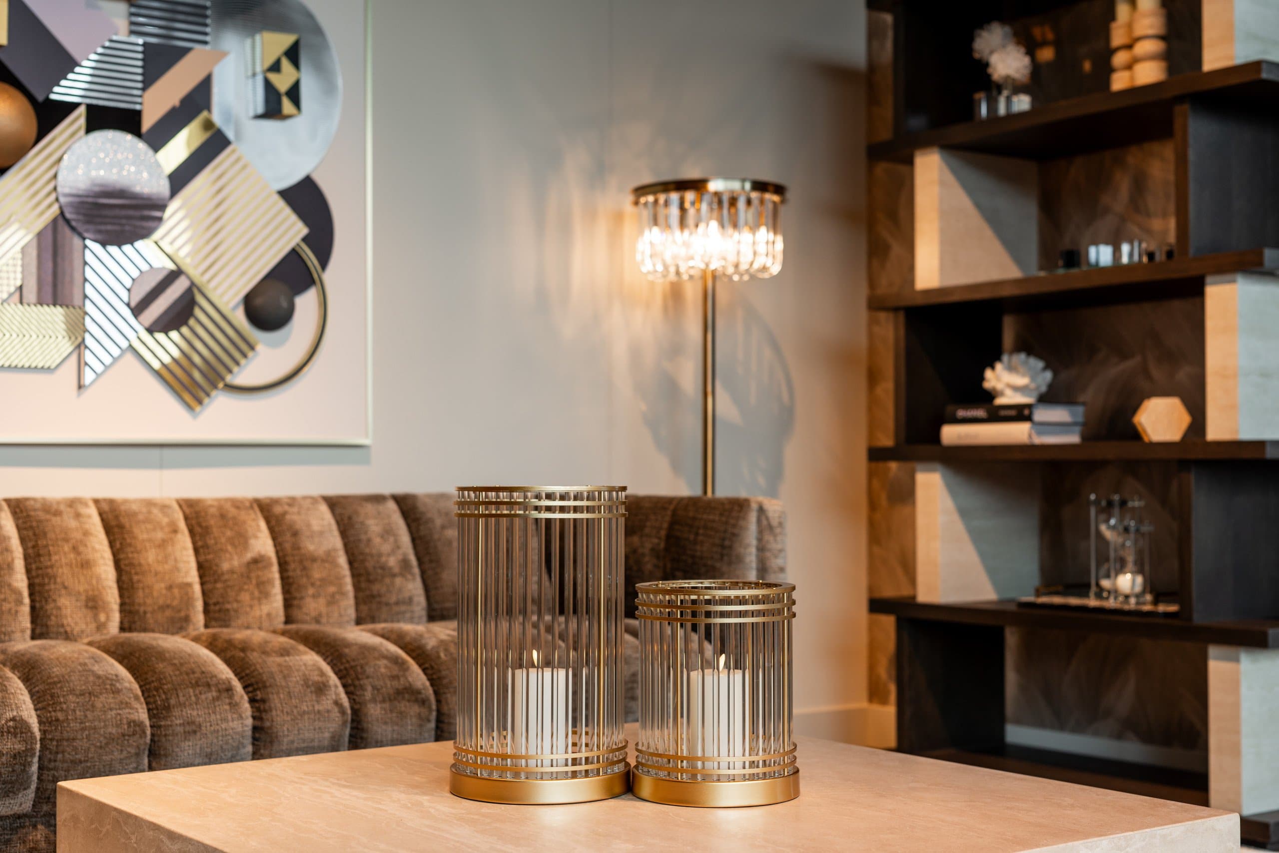 Verlichte vloerlamp met kristallen kap achter een bruine bank, met twee gouden windlichten met kaarsen op een tafel op de voorgrond