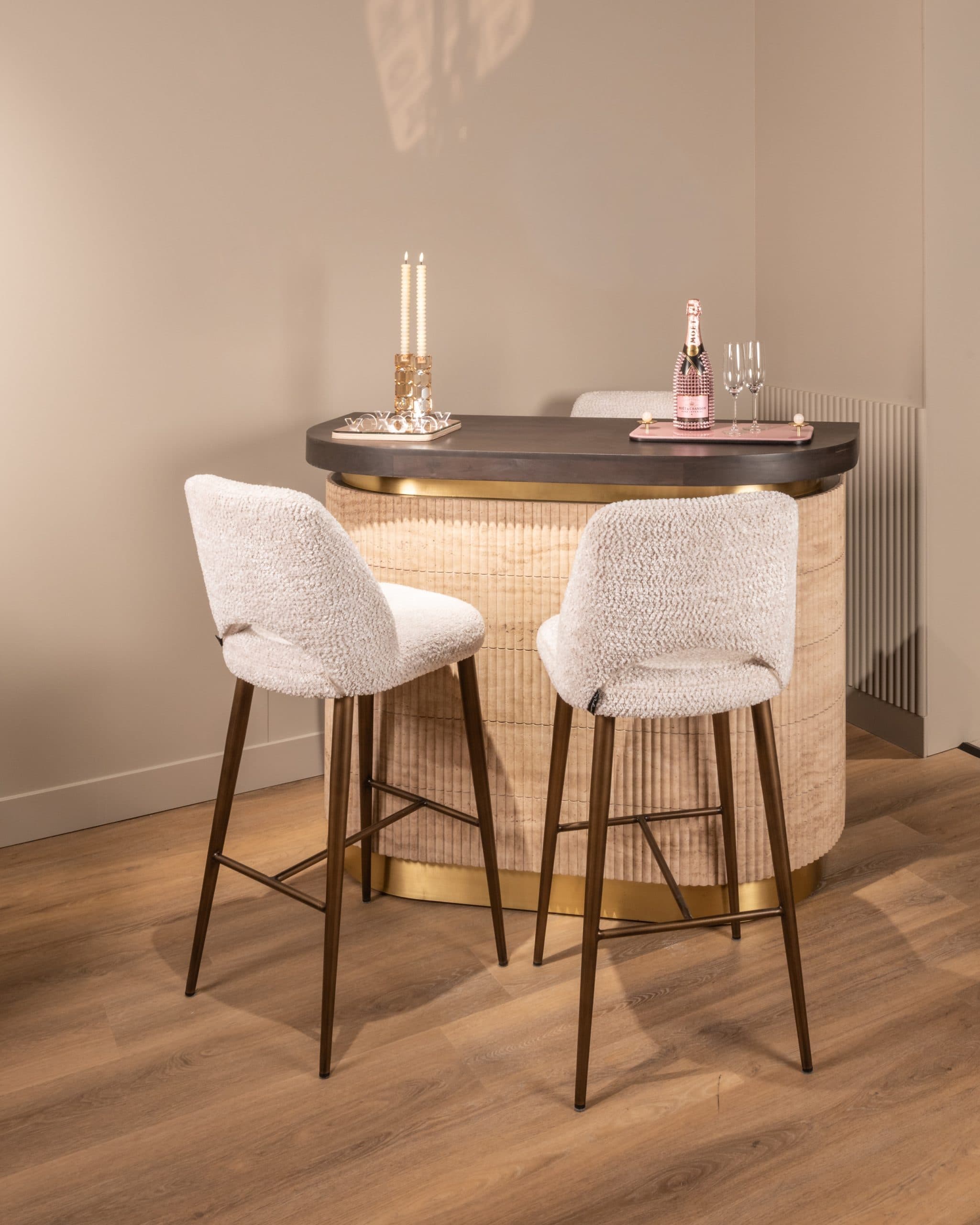 Ronde bar van geribd travertine met donker houten blad, twee bouclé barkrukken en een dienblad met glazen en een fles op het blad