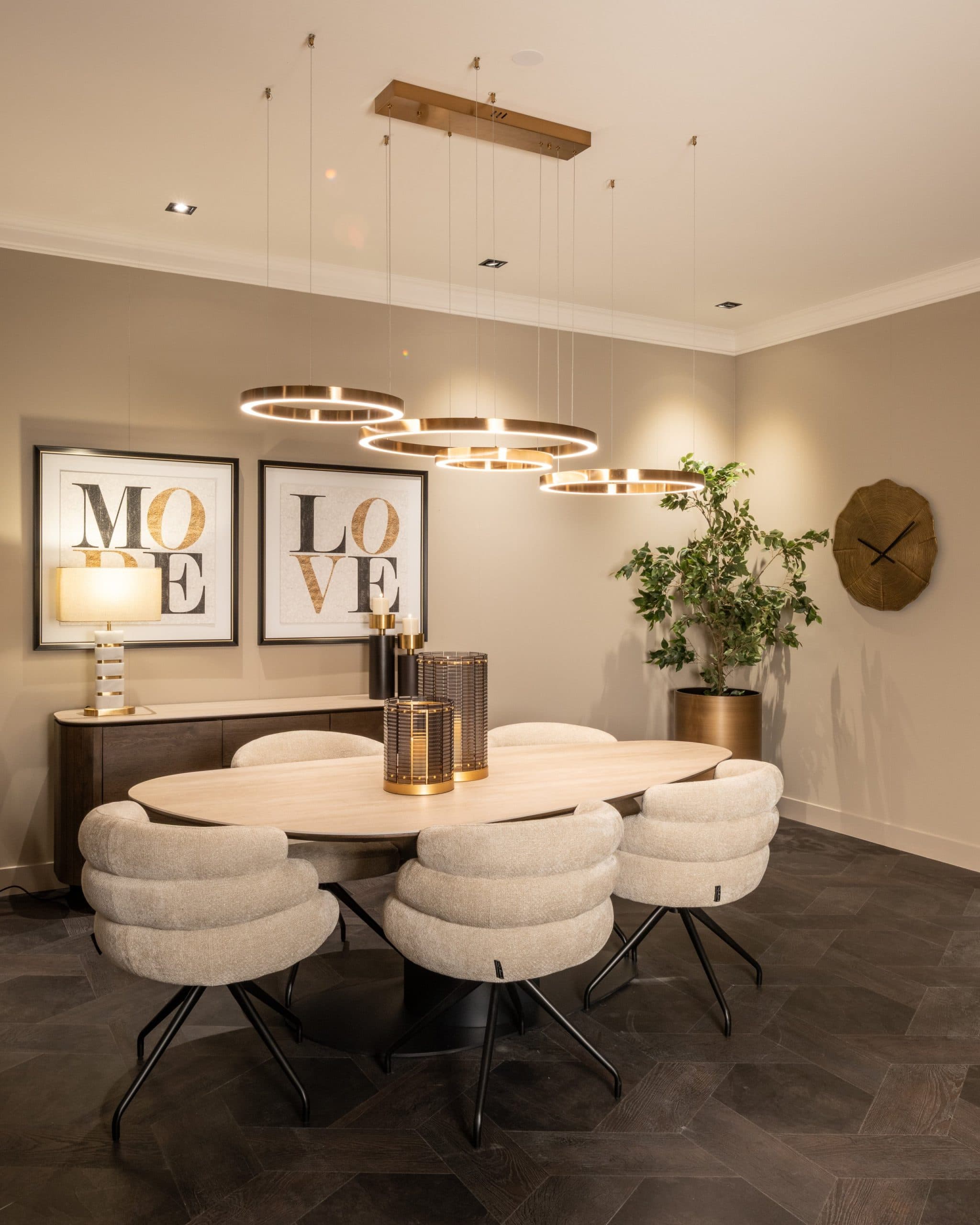 Ovale eettafel met beige gestoffeerde stoelen onder een hanglamp met vier gouden ringen, met dressoir, lijsten en een plant erachter.