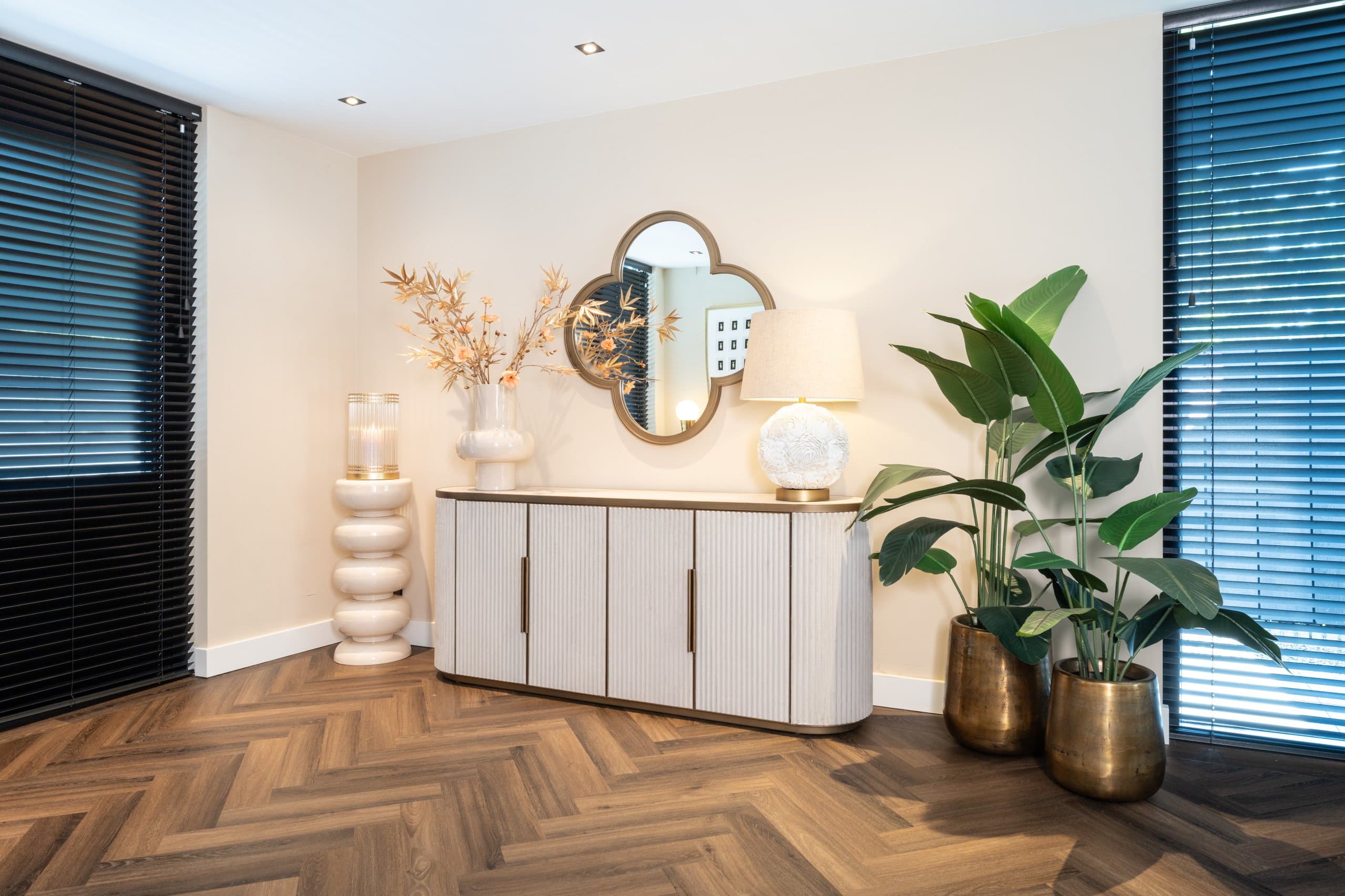 Off-white dressoir met geribbelde deuren, met daarboven een goudkleurige spiegel in klaverbladvorm, een tafellamp met witte kap, droogbloemen in een vaas en twee planten in messing potten.