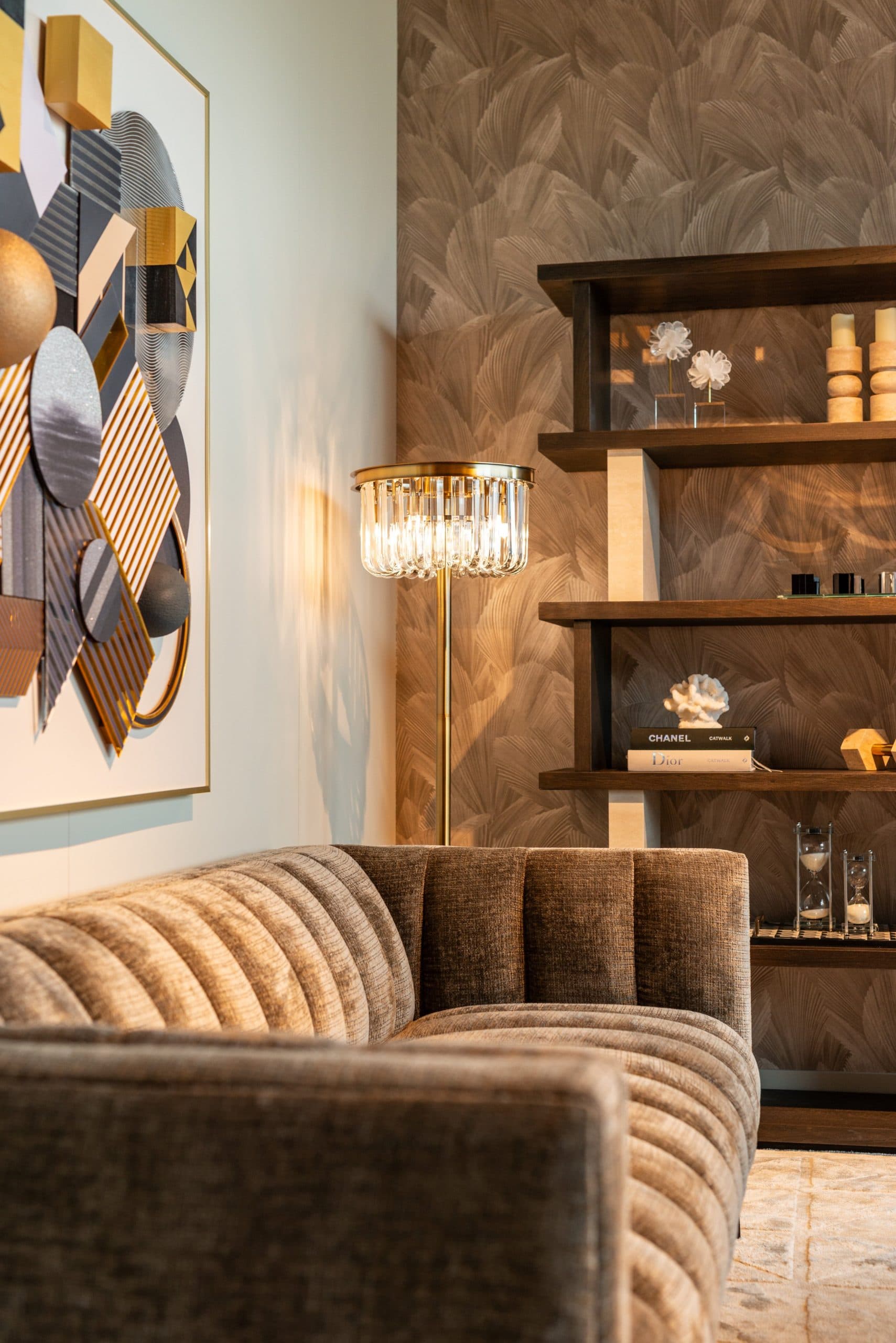 Woonkamerhoek met een gouden vloerlamp met kristallen kap die licht op een lichte wand werpt, naast een ingelijst kunstwerk, een houten wandpaneel en een bruine bank