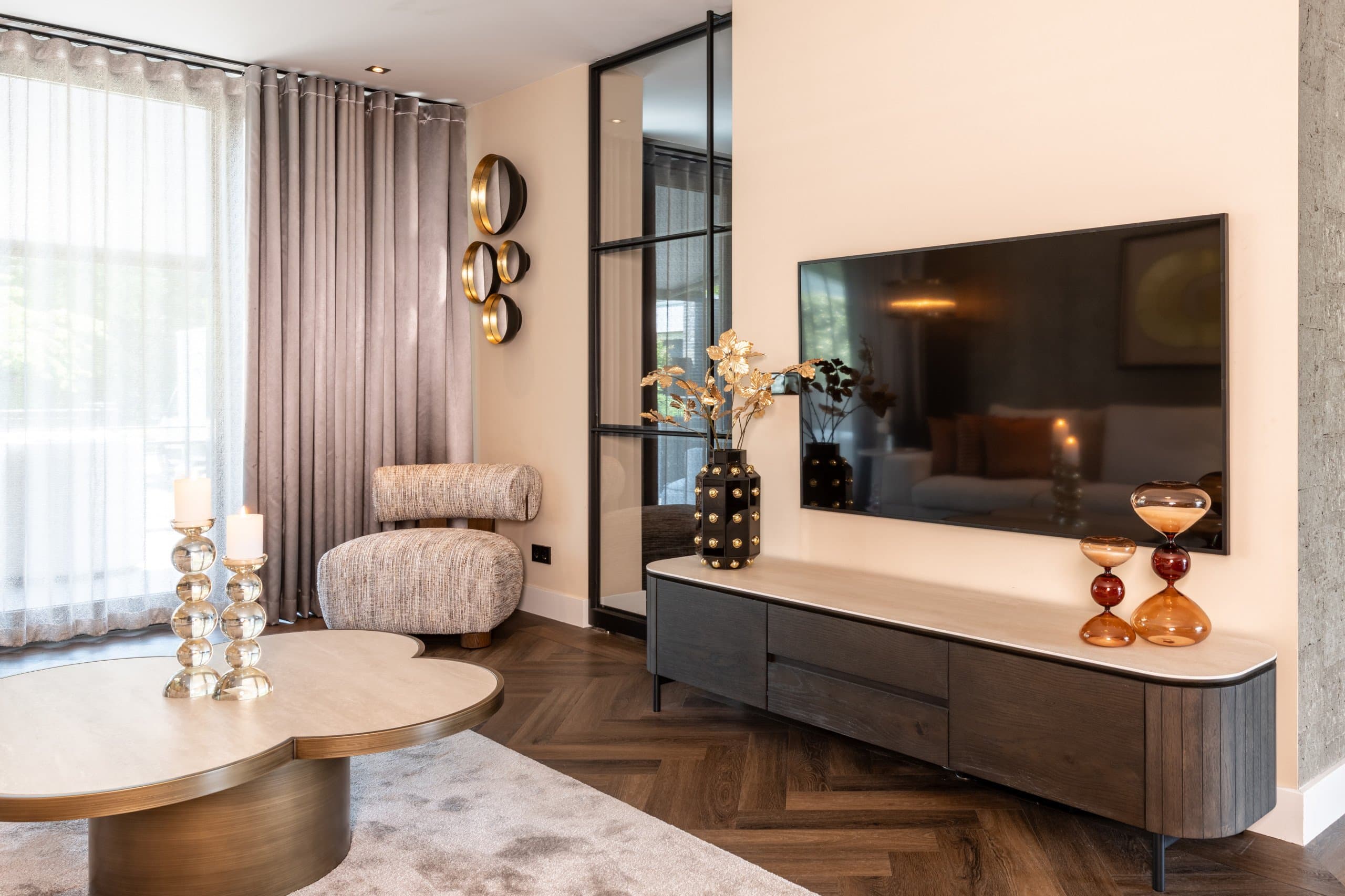 Woonkamer met een uitgeschakelde televisie aan een zandkleurige wand boven een donker tv-meubel met licht blad, met gordijnen en kaarsen op een lage tafel