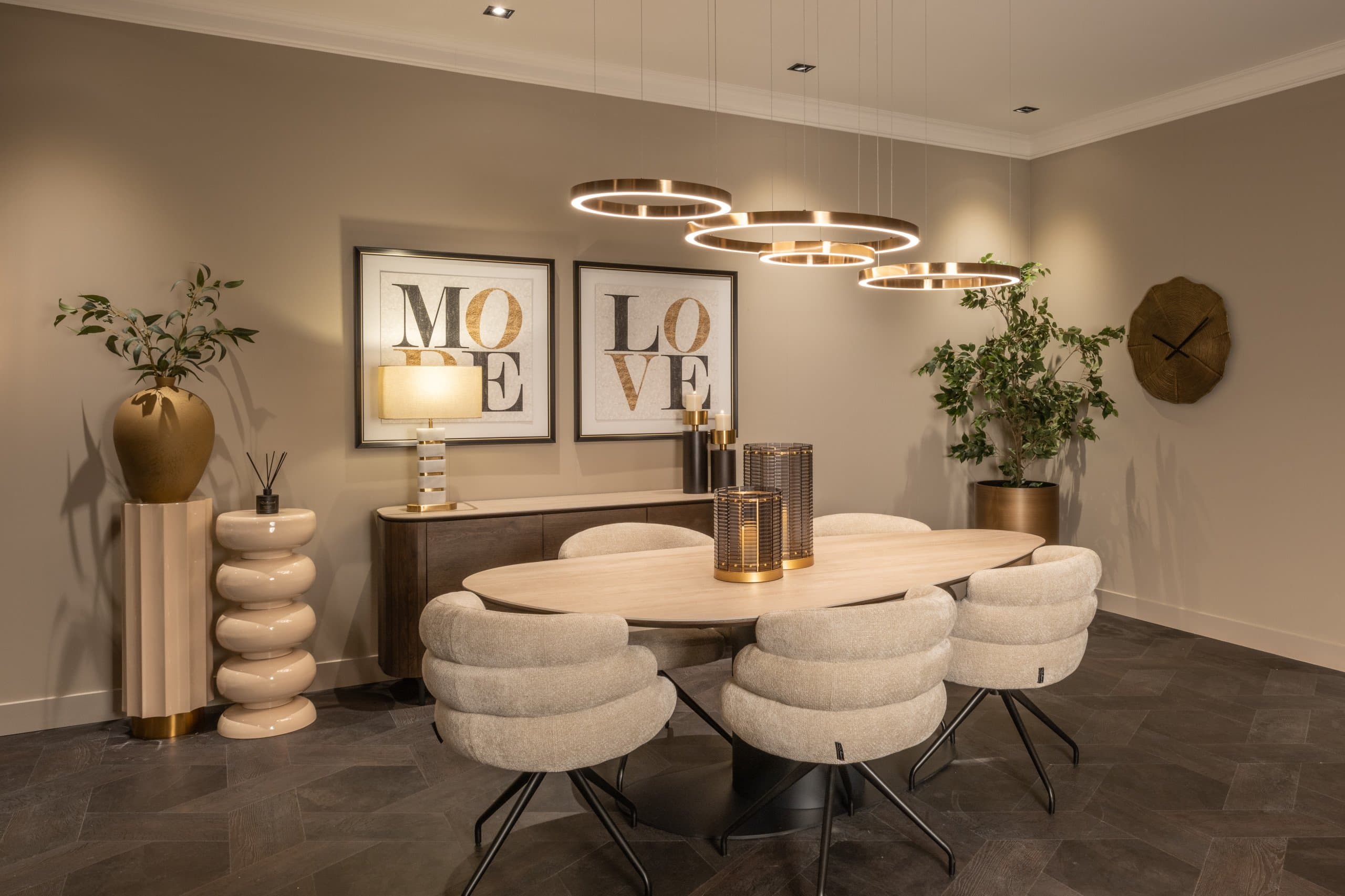 Eetkamer met een ovale lichte tafel, crèmekleurige stoelen, drie ringvormige hanglampen, een houten dressoir met twee ingelijste prenten en een hoge plant in de hoek