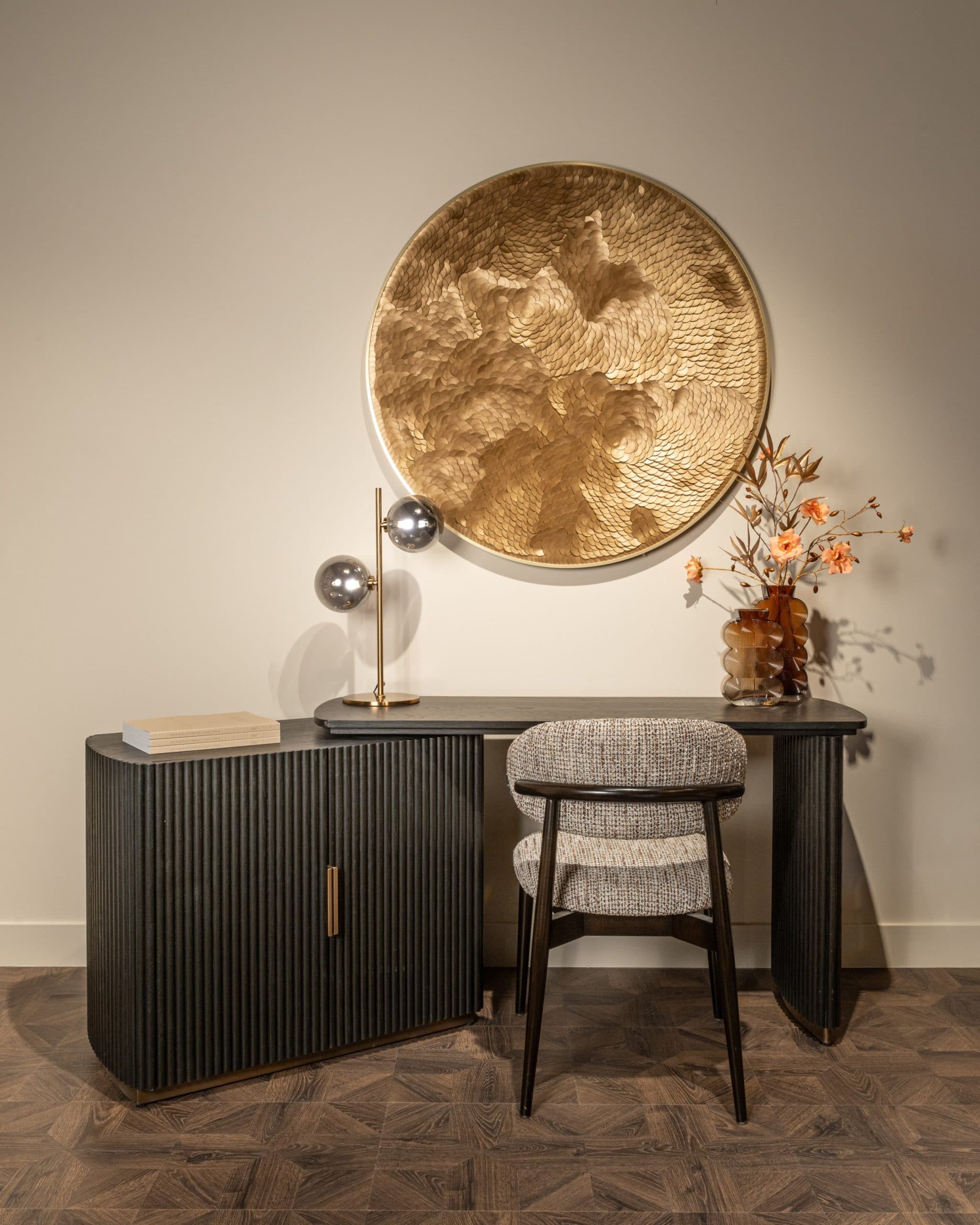 Tafellamp met twee rookglazen bollen op een donker geribbeld dressoir, met zachte schaduwen op de wand en een gouden wandobject erboven