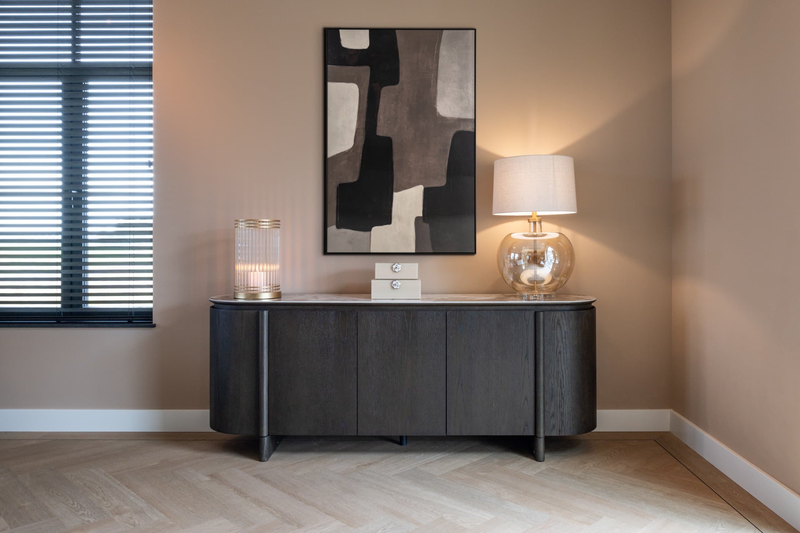 Donker dressoir tegen een warme wand met een brandende tafellamp met amberkleurige glazen voet, een windlicht en een abstract schilderij erboven