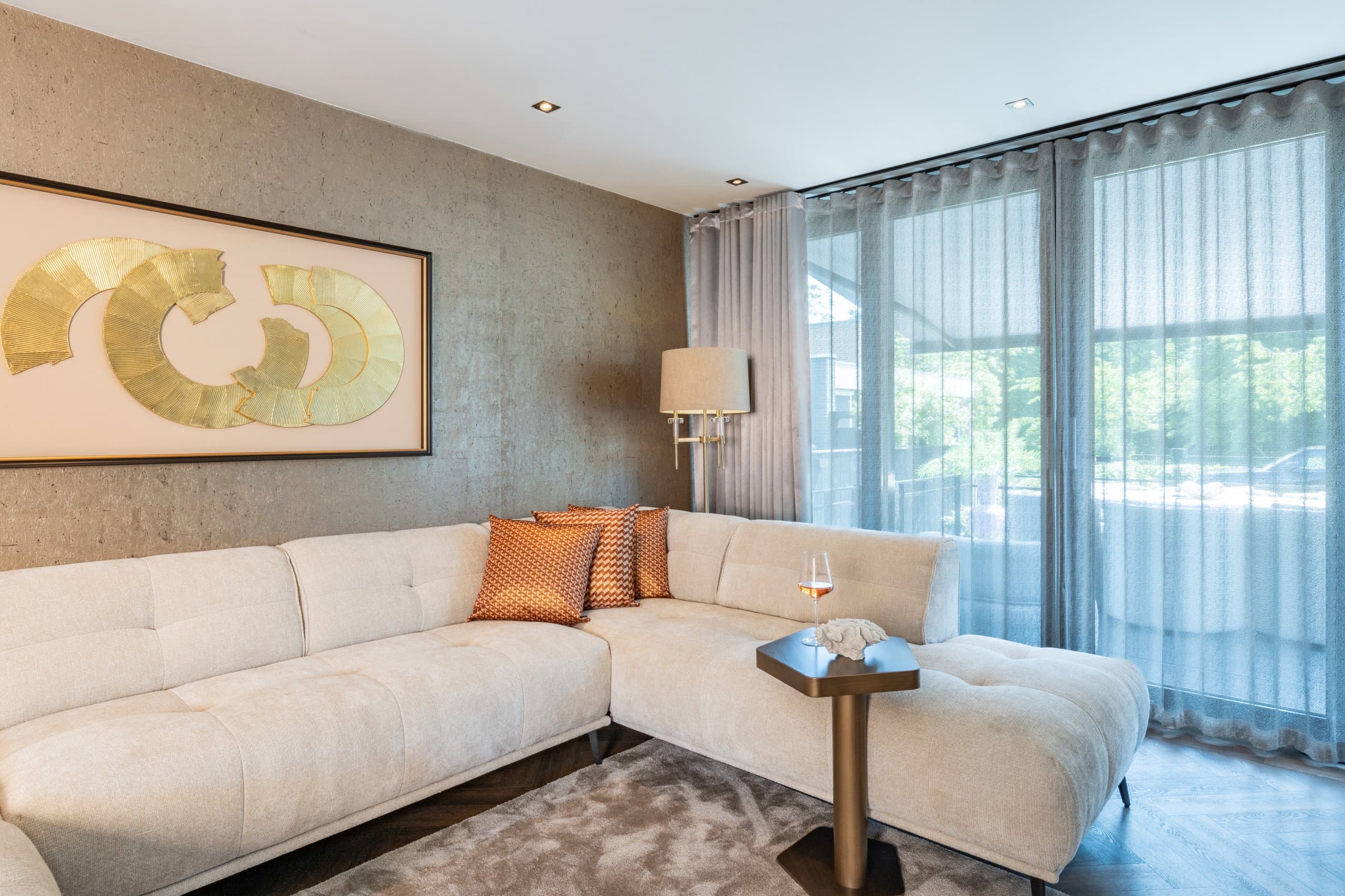 Woonkamer met crèmekleurige hoekbank, een wandlamp en daglicht dat door voile gordijnen naar binnen valt