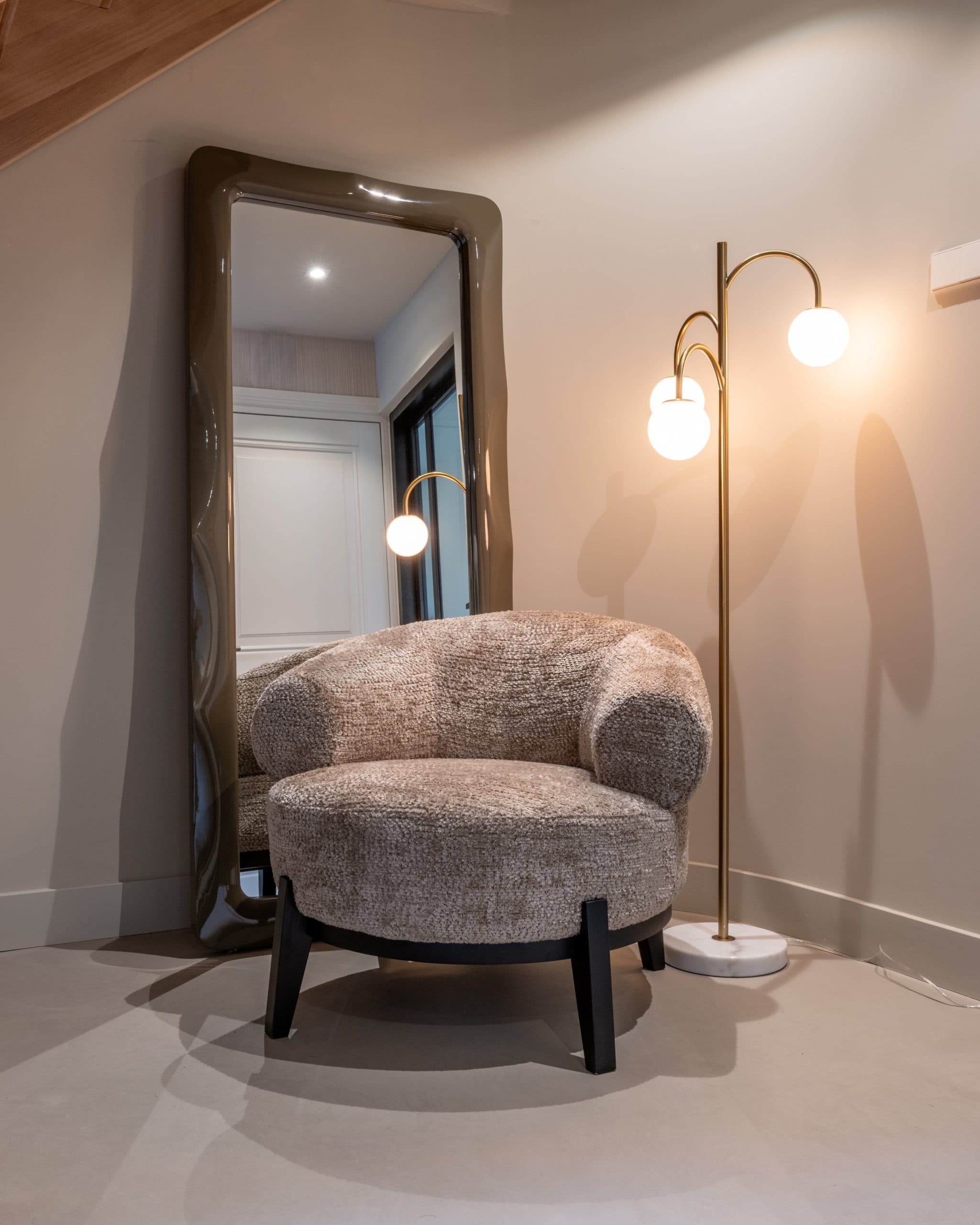 Goudkleurige vloerlamp met witte glazen bollen naast een ronde beige bouclé fauteuil, met een grote spiegel op de achtergrond