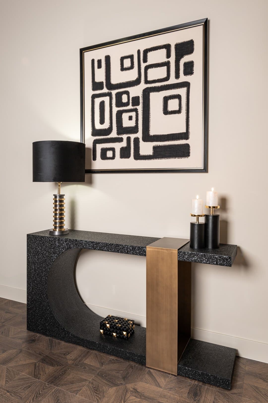 Zwarte terrazzo console tegen een beige wand met een tafellamp met zwarte kap, twee kaarsen op zwarte houders en een abstract zwart-wit schilderij erboven