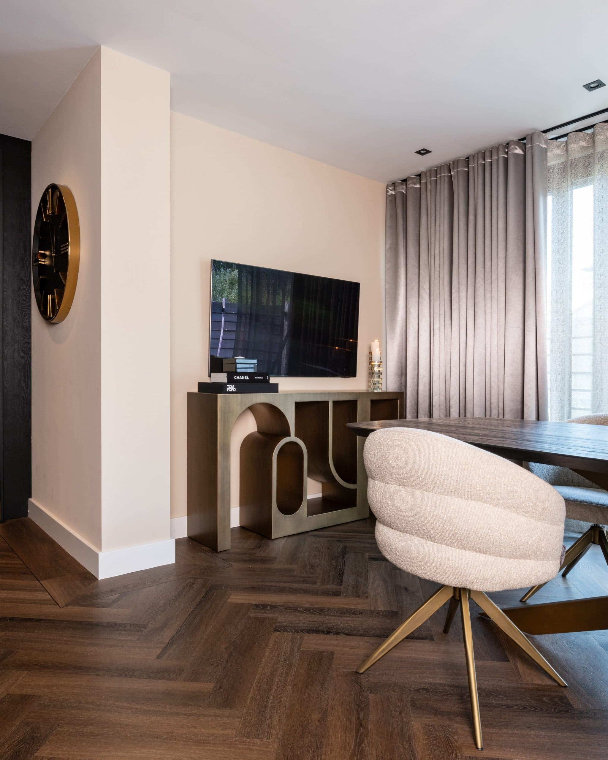 Woonkamerhoek met een televisie op een goudkleurige console, een ronde klok aan de muur, grijze gordijnen en een lichte eetkamerstoel