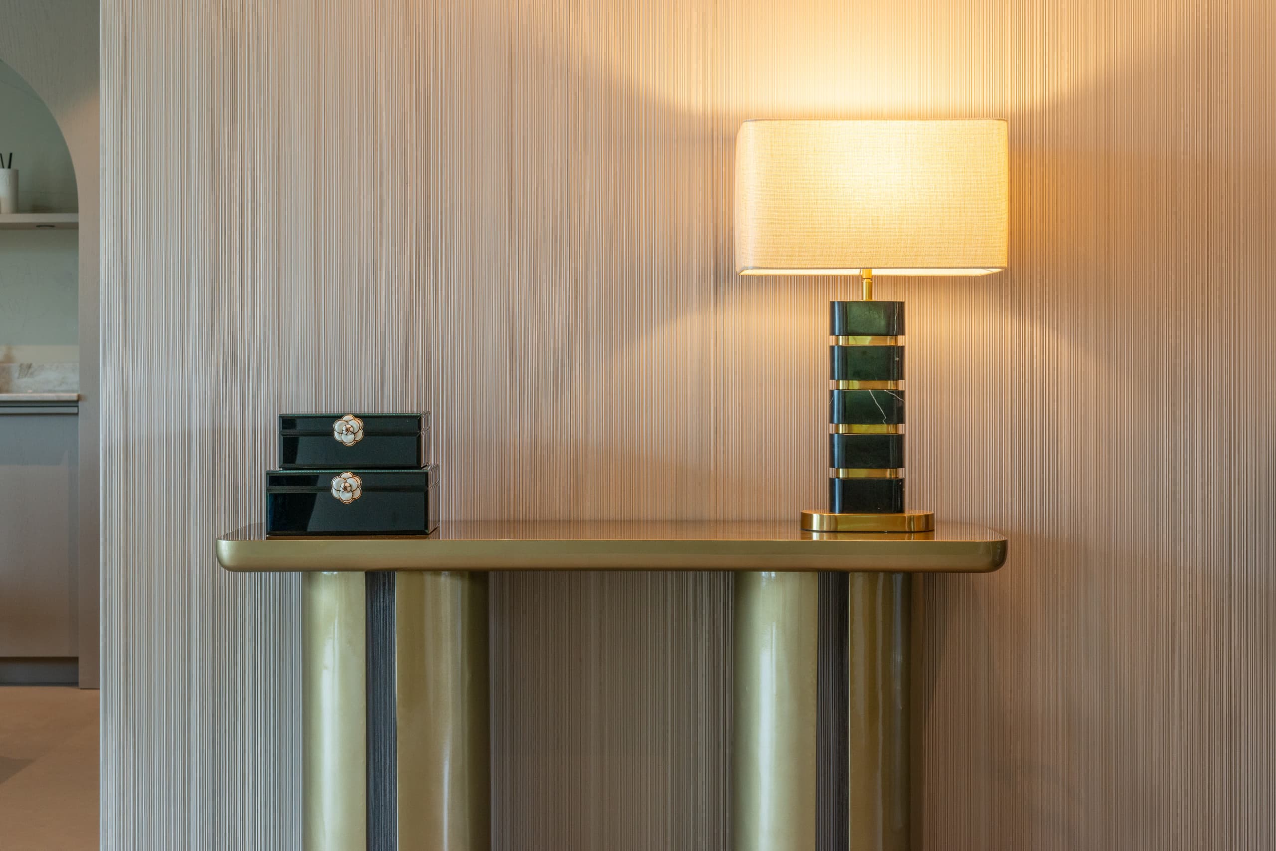 Goudkleurige consoletafel tegen een geribde wand, met een tafellamp op een groen marmeren voet en twee zwarte lakdozen ernaast