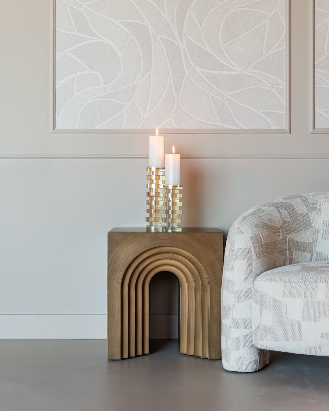 Twee kandelaars met een goudkleurig vlechtpatroon en brandende stompkaarsen op een houten sidetable, naast een fauteuil met gedessineerde stof