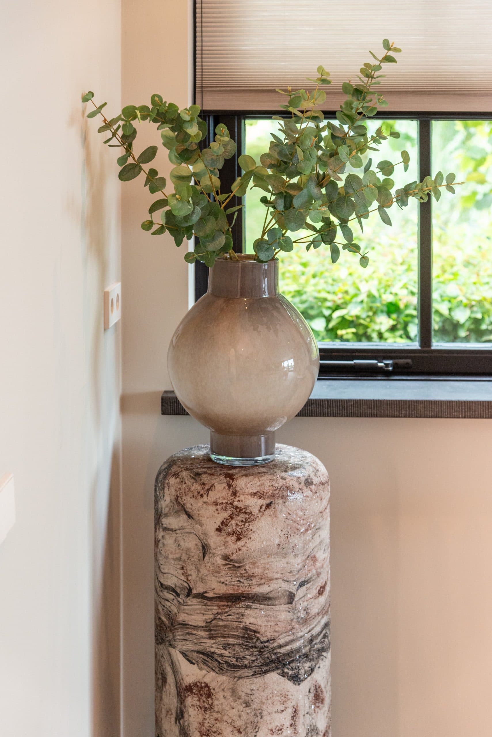 Taupe glazen bolvaas met eucalyptustakken op een ruwe stenen sokkel, voor een raam met zwart kozijn en groen buiten