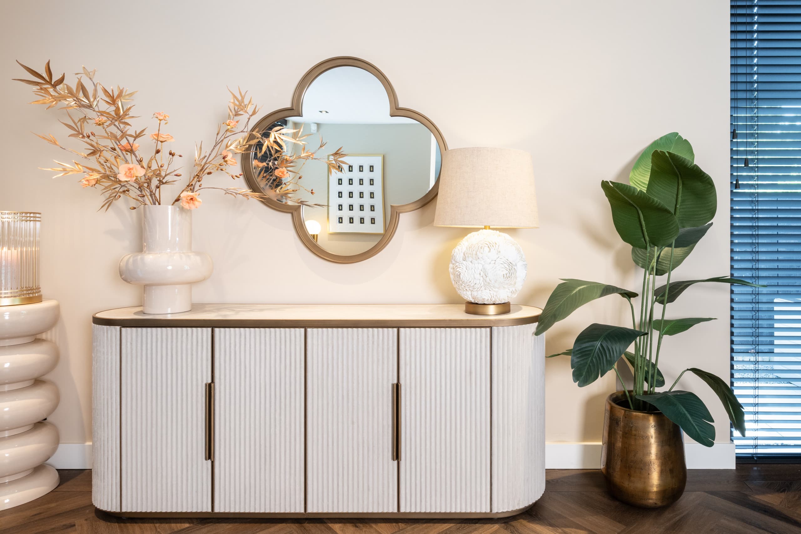 Wandspiegel met gouden klaverbladvorm boven een licht ribbeldressoir, met een ronde tafellamp met linnen kap ernaast en een plant in een bronzen pot