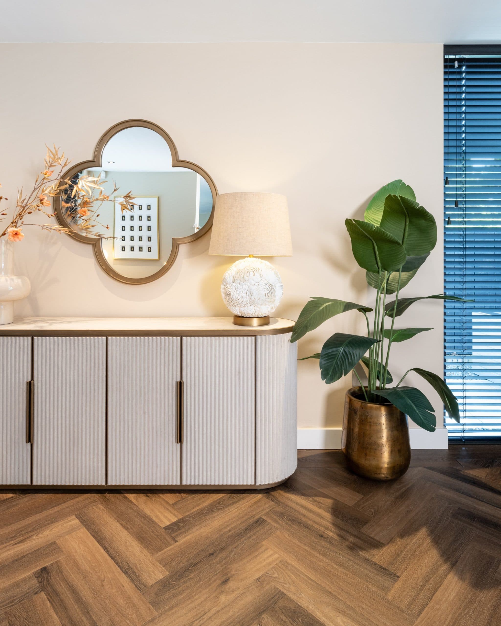 Gouden wandspiegel boven een licht geribbeld dressoir met een tafellamp met beige kap en een grote groene plant ernaast.