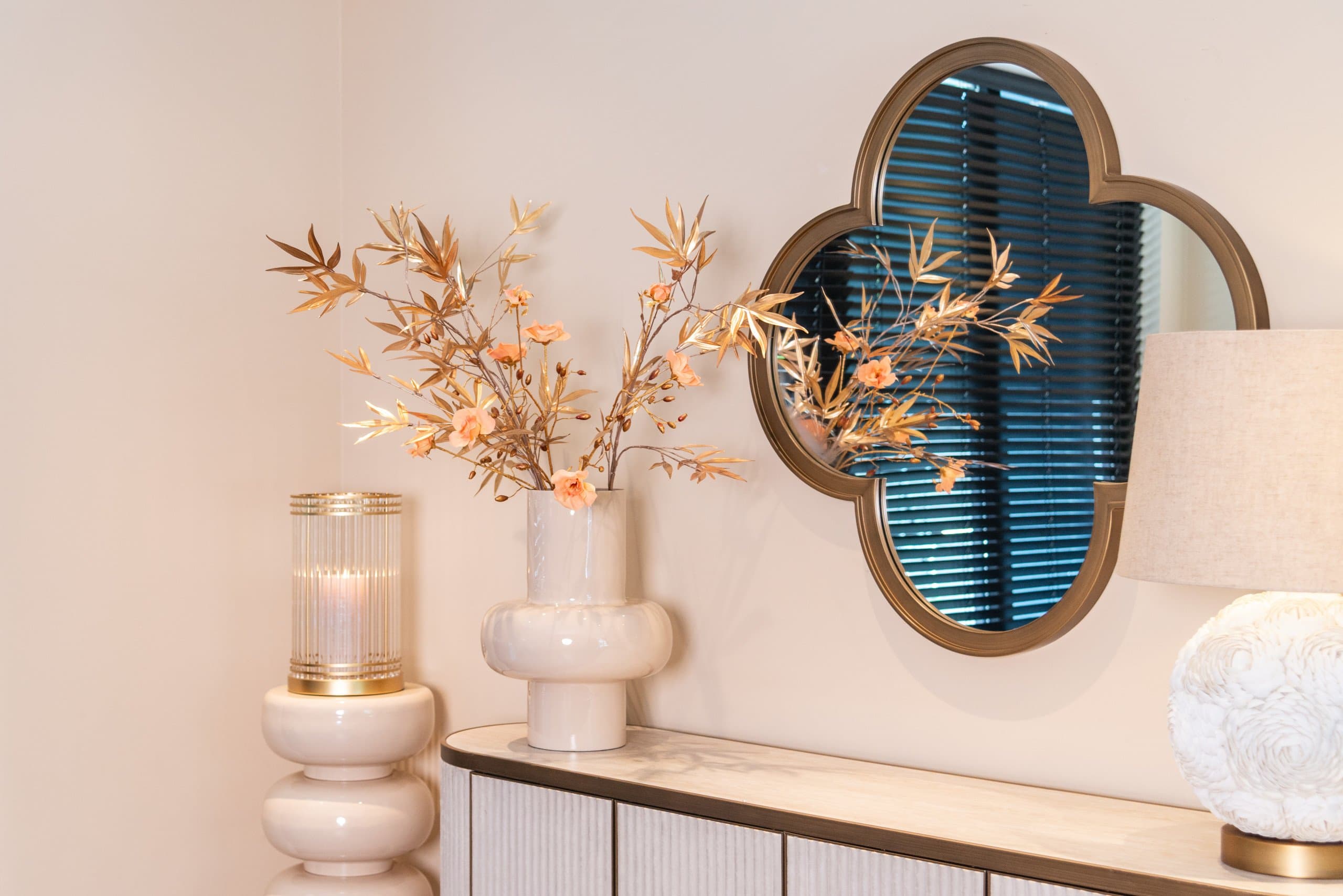 Hoek van een kamer met een goudkleurige wandspiegel, een witte vaas met gouden takken en een glazen windlicht op een donker meubelblad