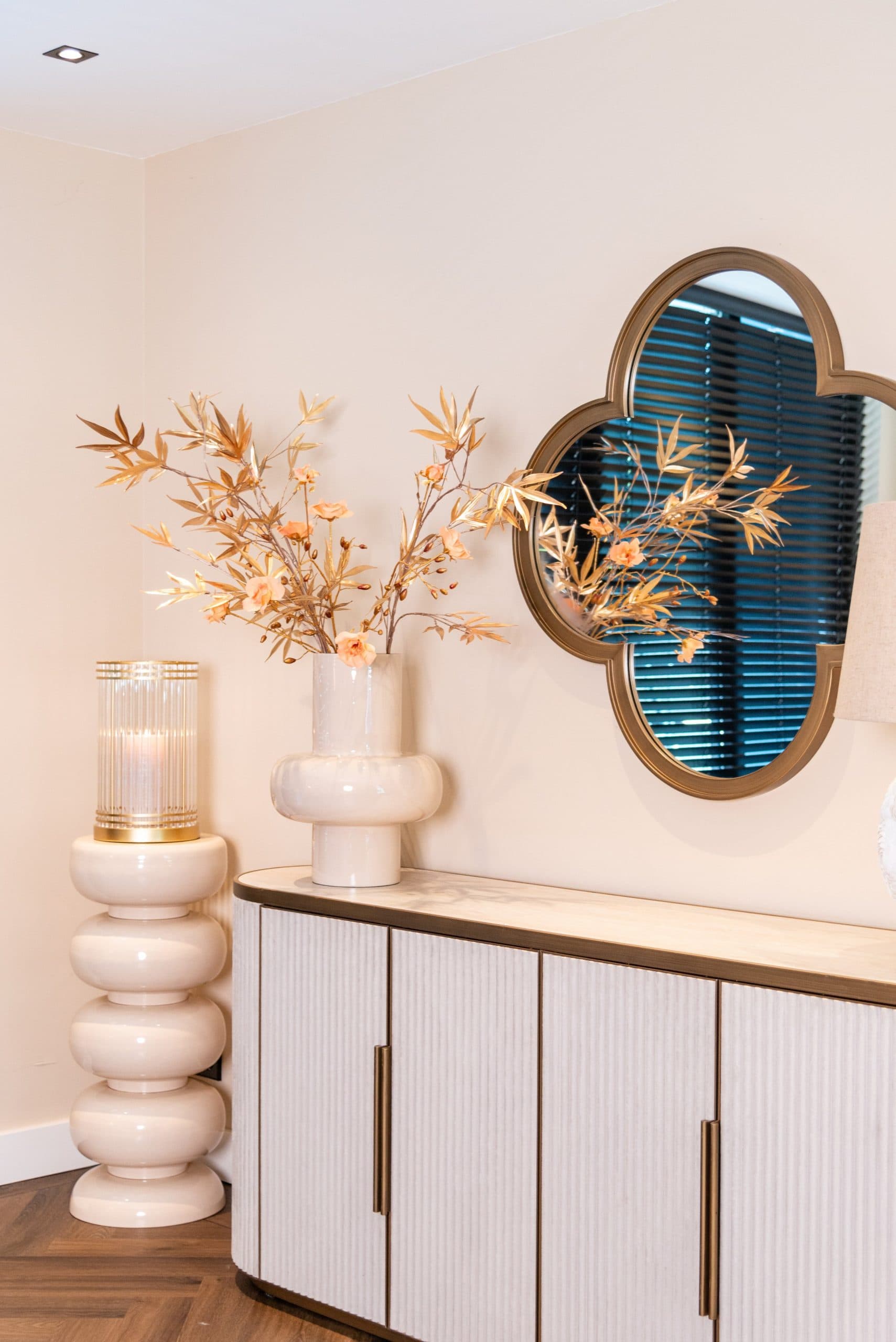 Consoletafel met een brandend windlicht in geribbeld glas, een crèmekleurige vaas met gouden takken en een gouden spiegel die het raam erachter weerkaatst