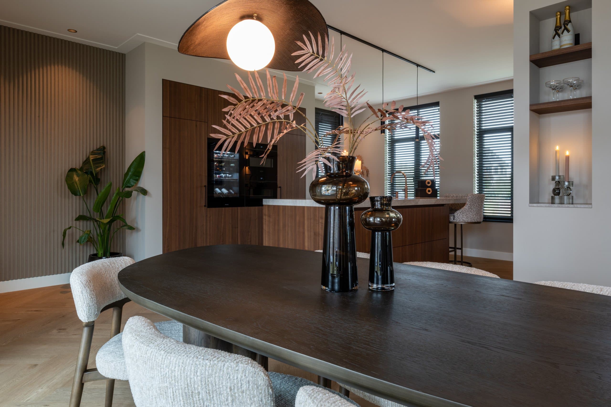 Ovale eettafel met donker blad en twee rookglazen vazen met roze palmtakken als middenstuk, omringd door witte bouclé stoelen