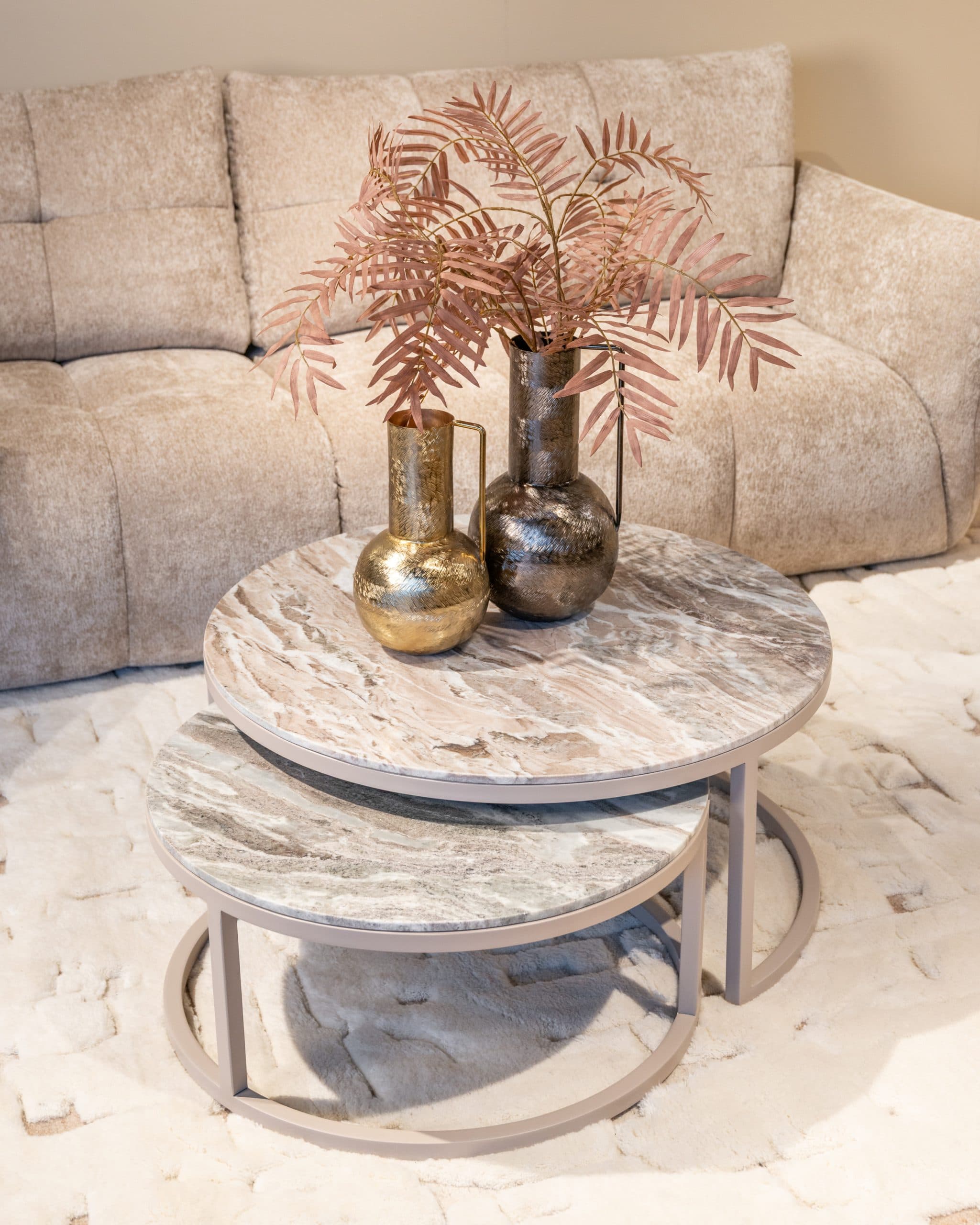 Twee ronde salontafels met marmeren blad en gouden vazen met roze takken, voor een beige geribbelde bank op een licht vloerkleed