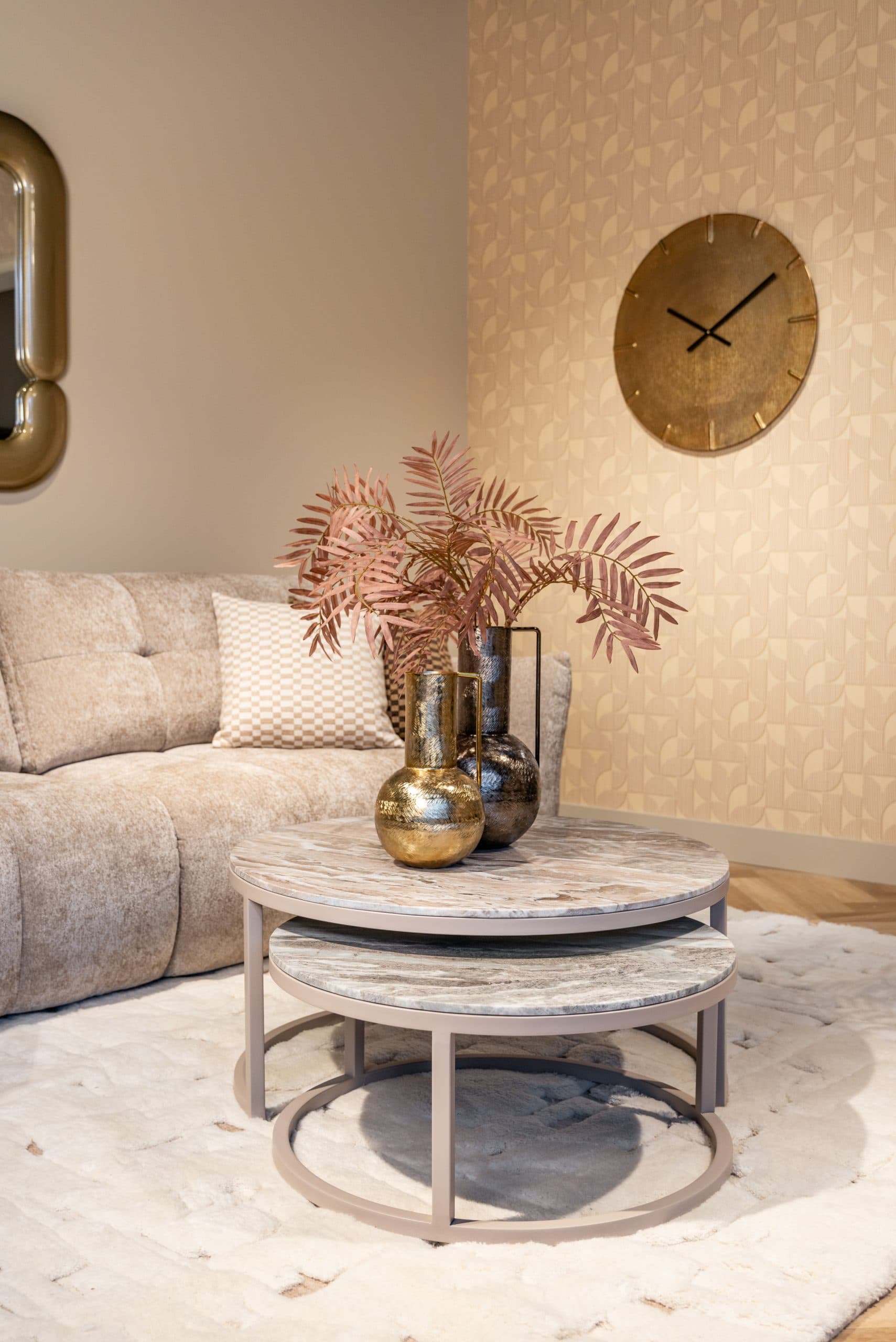 Hoek van een woonkamer met een beige bank, een geblokt sierkussen en twee metalen vazen met een roze varentak op een ronde bijzettafel met marmerlook