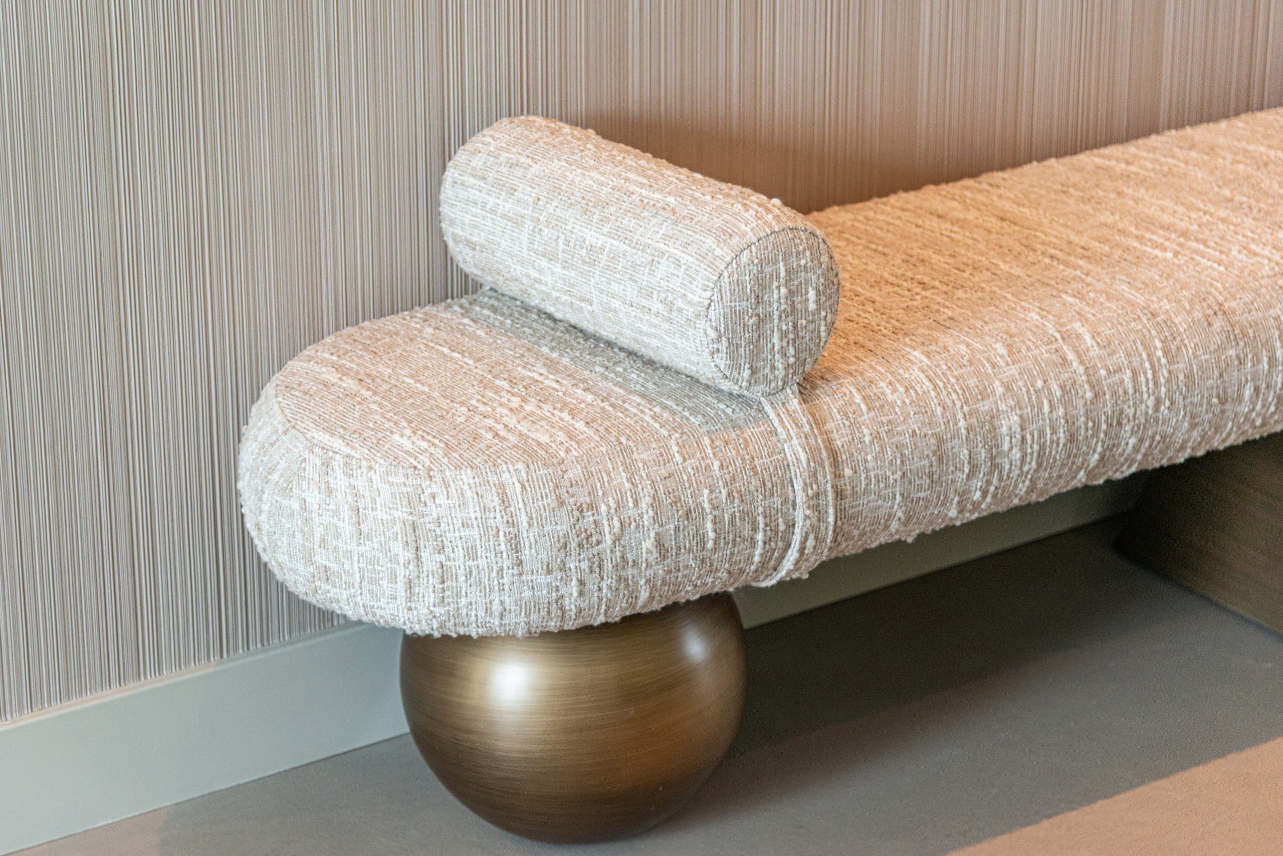 Detail van een crèmekleurige bouclé bank met zichtbare lusstructuur, een ronde rolkussen en een bronskleurige bolpoot, tegen een lichte geribbelde wand