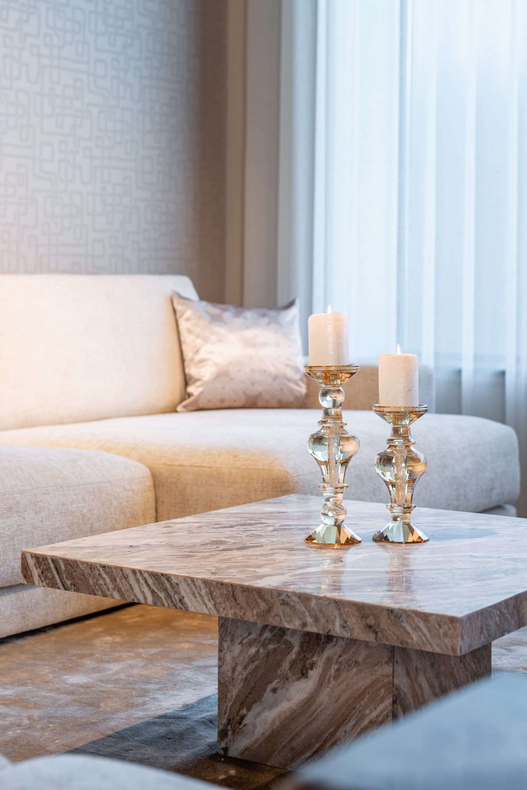 Twee brandende stompkaarsen op glazen kandelaars van verschillende hoogte op een marmeren salontafel, met daarachter een crèmekleurige bank en een gedessineerd sierkussen.