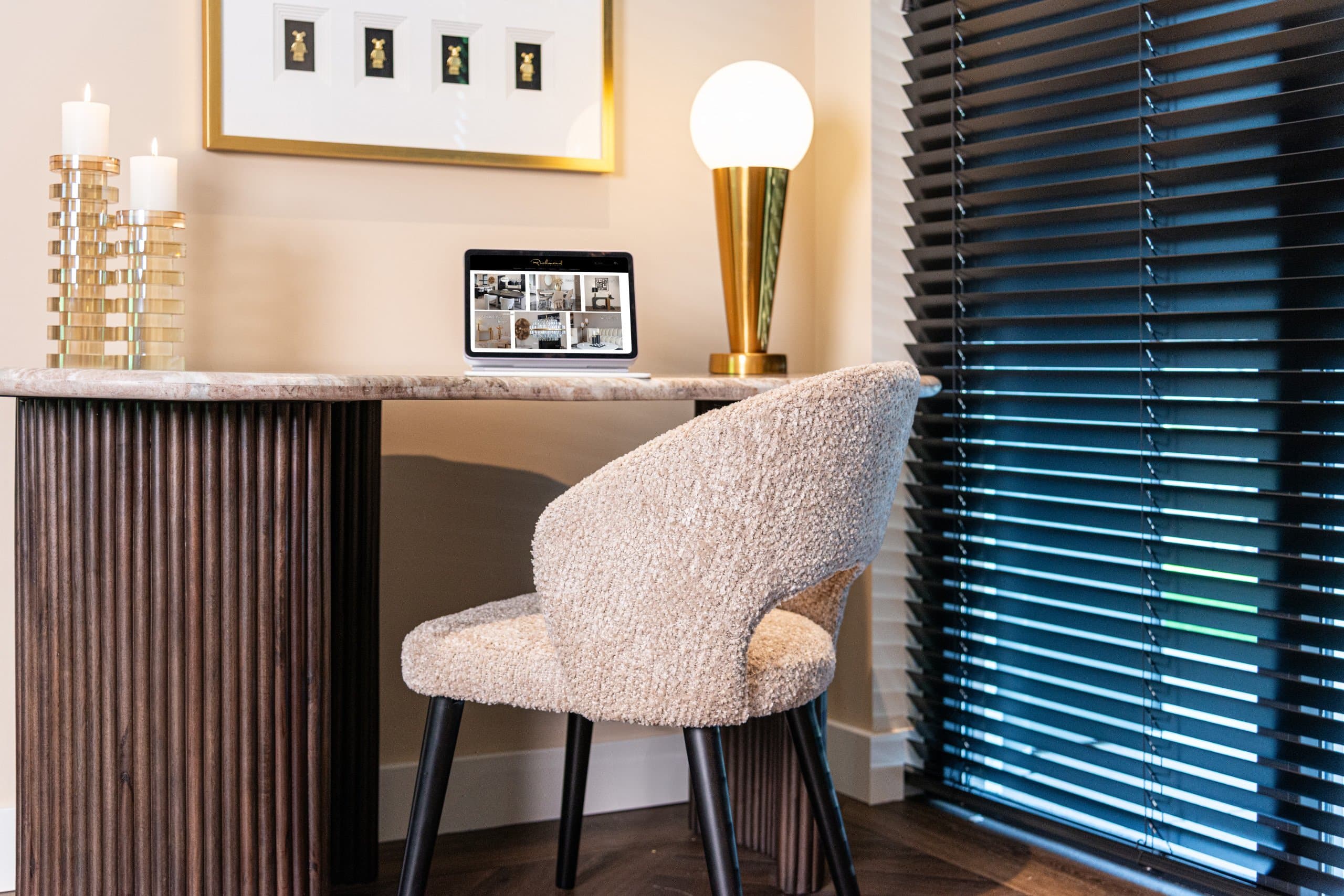 Werkplek met een boucle stoel aan een geribbeld bureau bij een raam met donkere horizontale jaloezieën, met een goudkleurige tafellamp en twee kandelaars met witte kaarsen