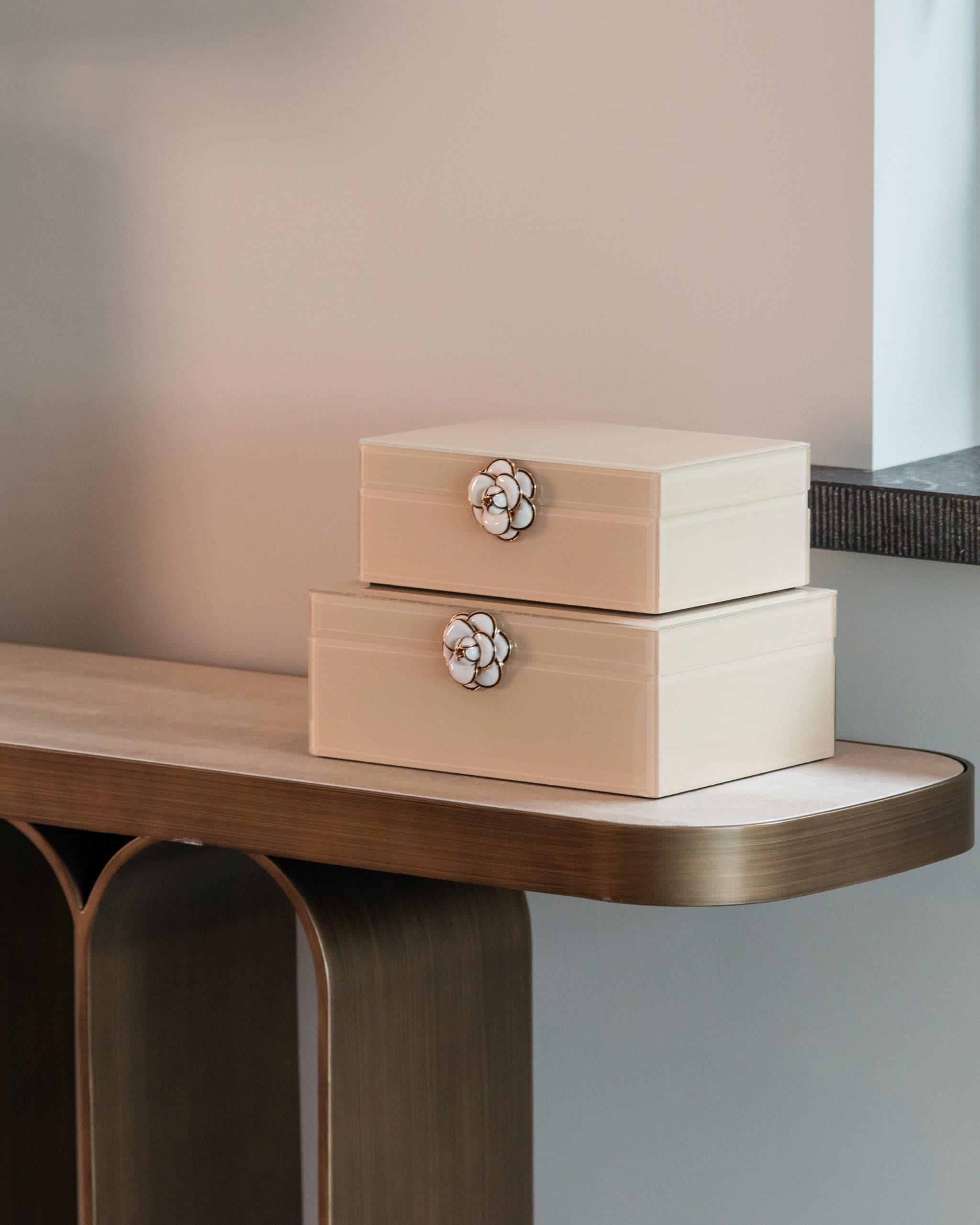 Twee gestapelde ivoorkleurige opbergdozen met een bloemvormige sluiting op een houten console tegen een zachtroze wand
