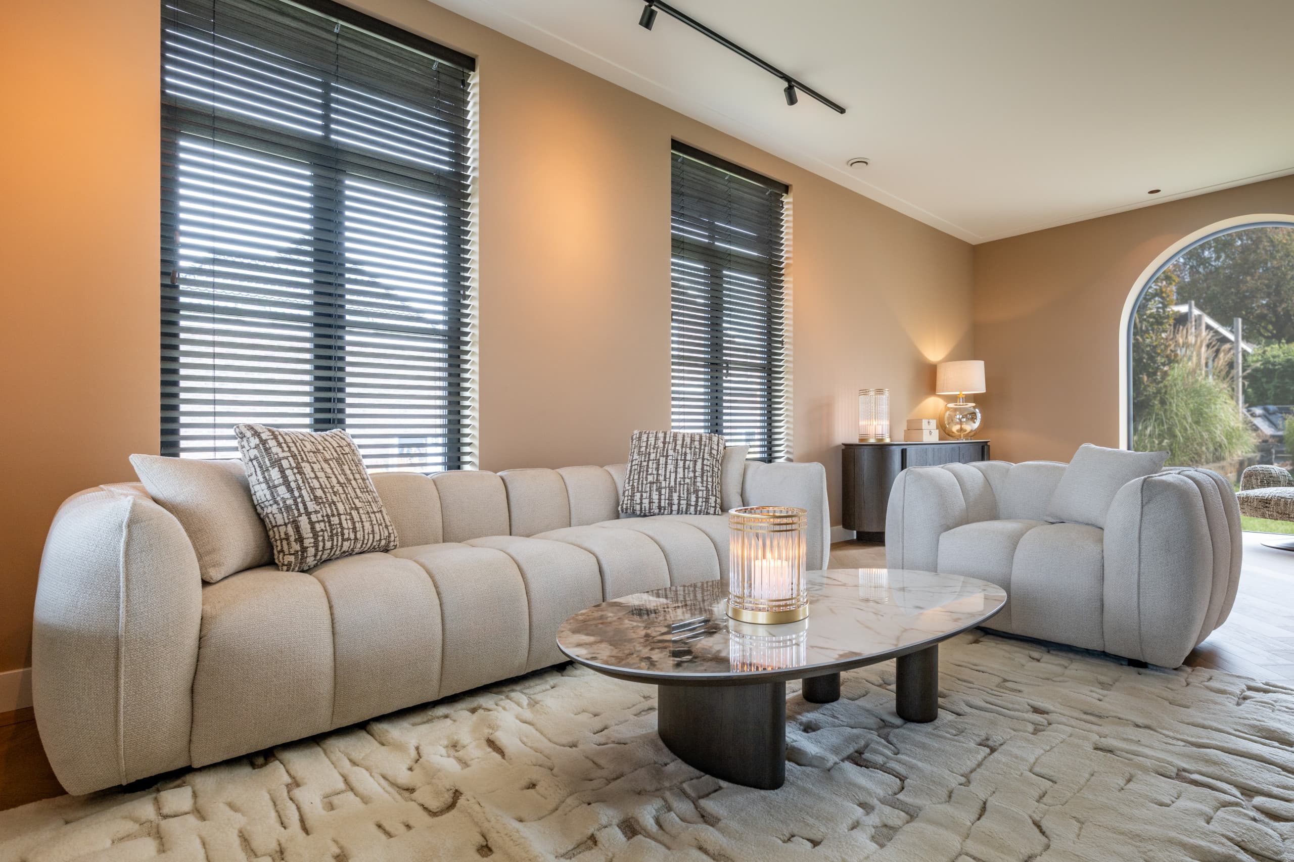 Woonkamer met hoge ramen met donkere jaloezieën, een crèmekleurige geribbelde bank met sierkussens, een ronde fauteuil en een groot licht vloerkleed