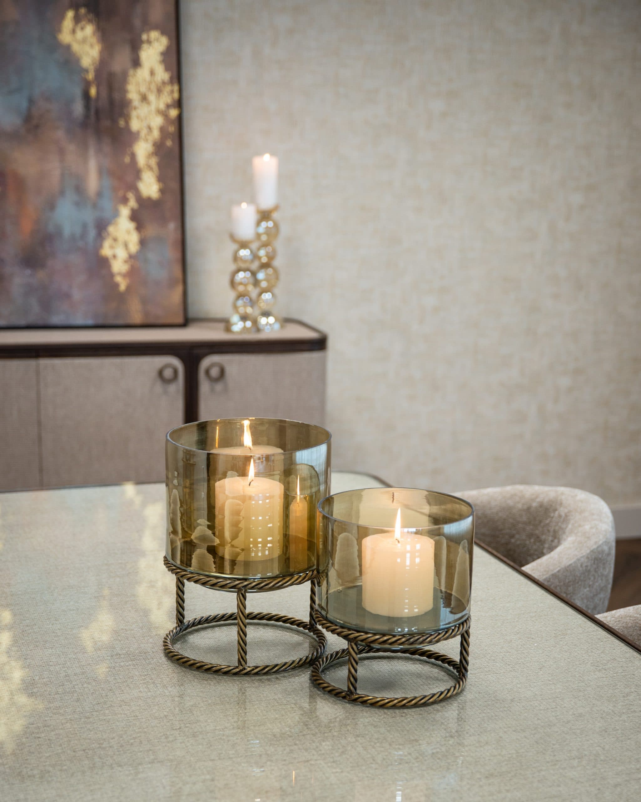 Twee glazen windlichten in rookglas met brandende stompkaarsen op een tafel, met op de achtergrond een dressoir met linnen fronten