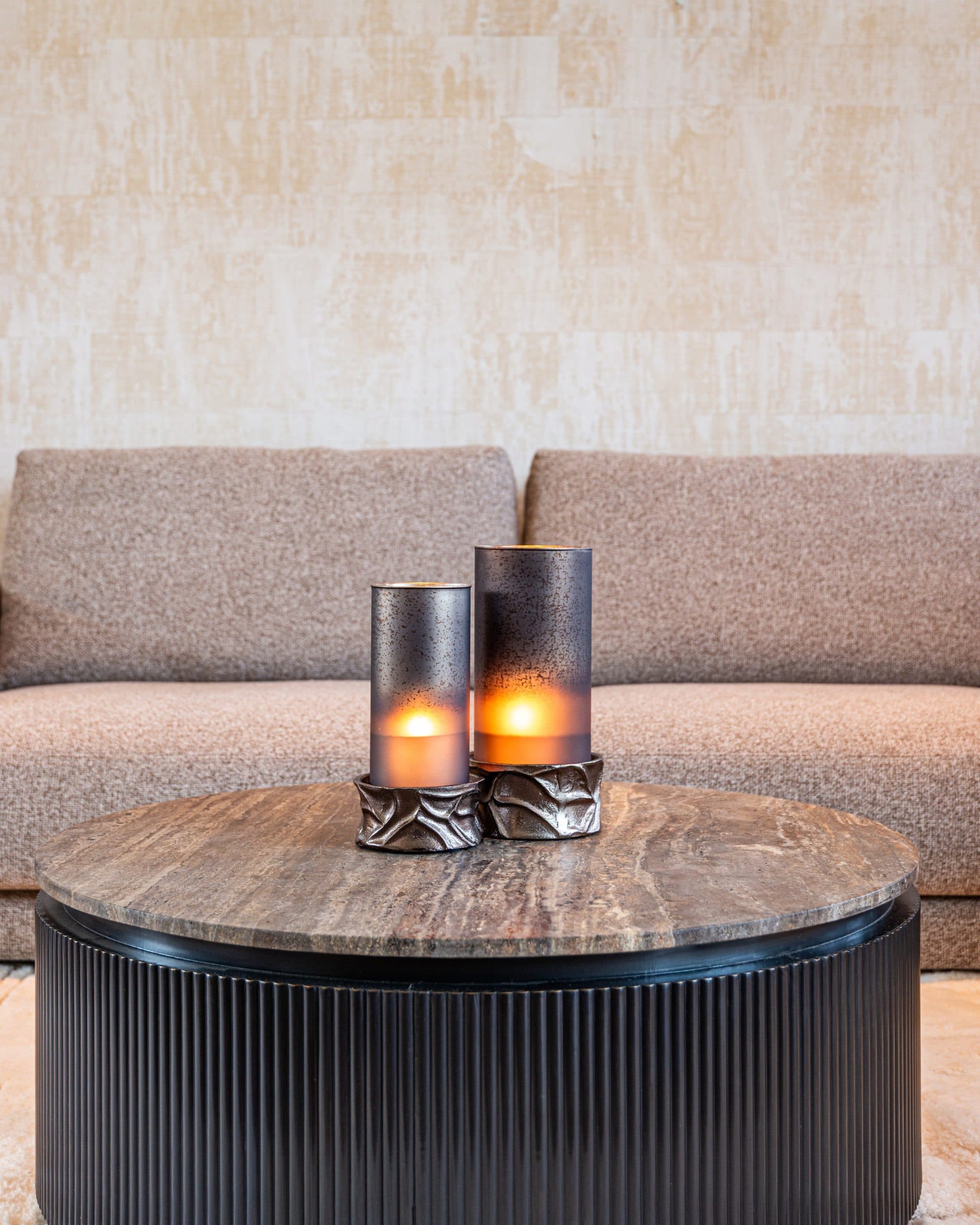 Twee smoke glazen windlichten met brandende kaarsen op een ronde travertijnen tafel voor een beige stoffen bank