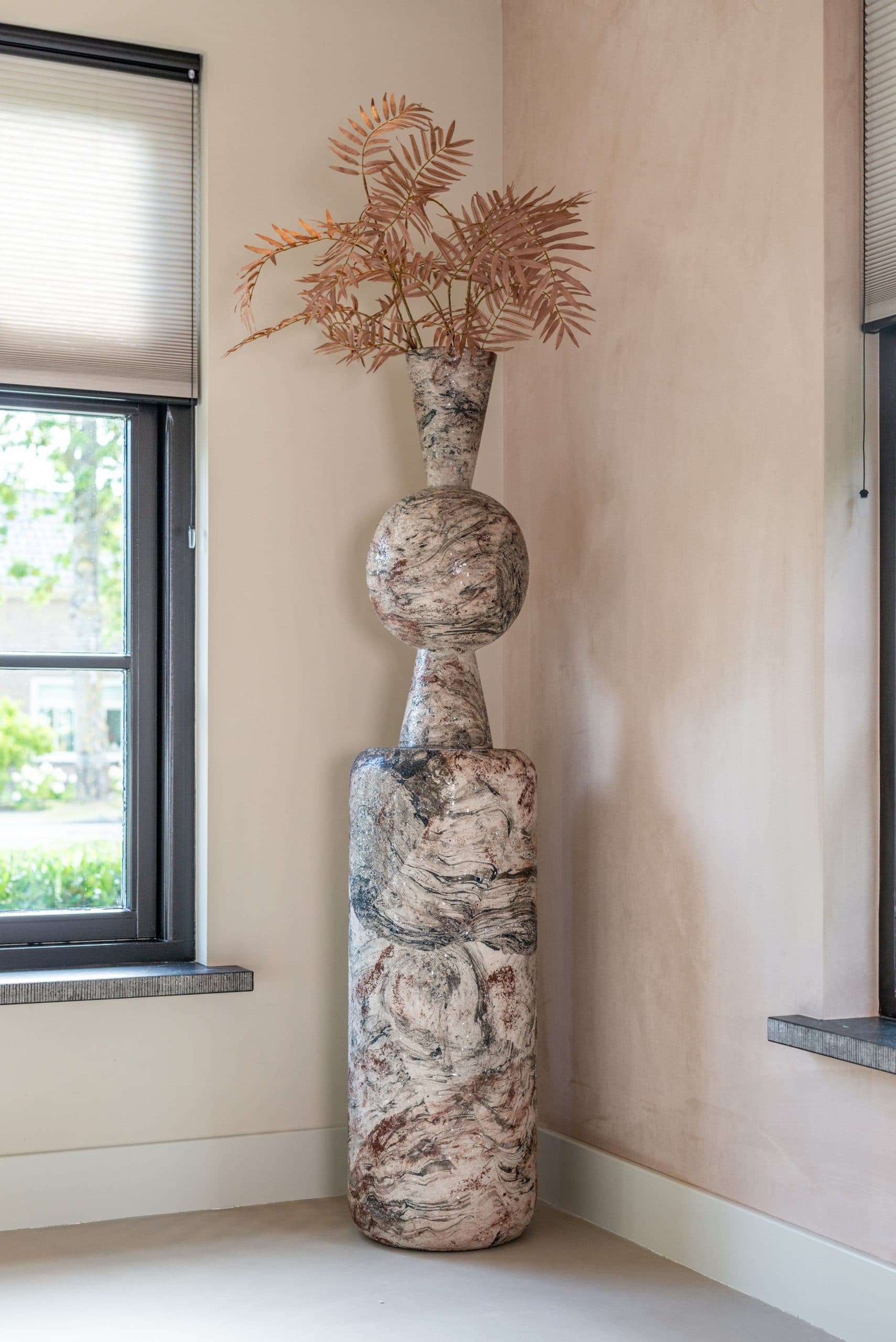 Hoge pilaar met beige-bruine marmerlook in een hoek naast een raam, met bovenop een sculpturale vaas met bolvorm