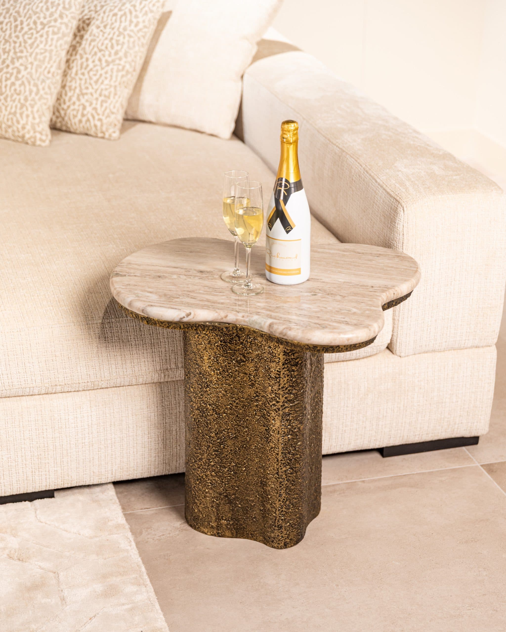 Bijzettafel met een organisch gevormd travertine blad en een grofgestructureerde bronskleurige voet, deels onder een crèmekleurige bank geschoven