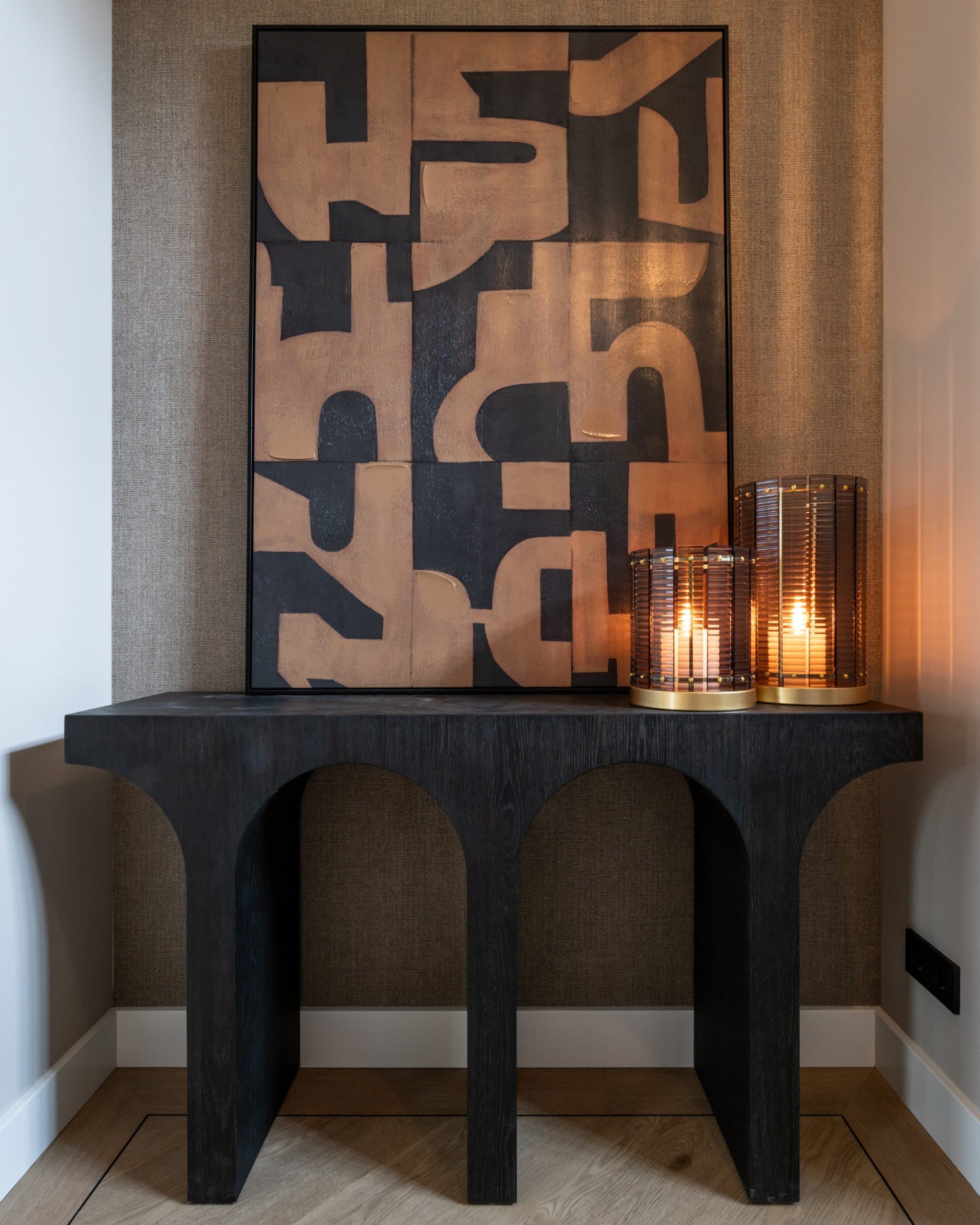 Wandvlak bekleed met grof linnen tussen twee witte muren, met daarvoor een zwarte console met boogpoten, een grafisch canvas en twee brandende windlichten