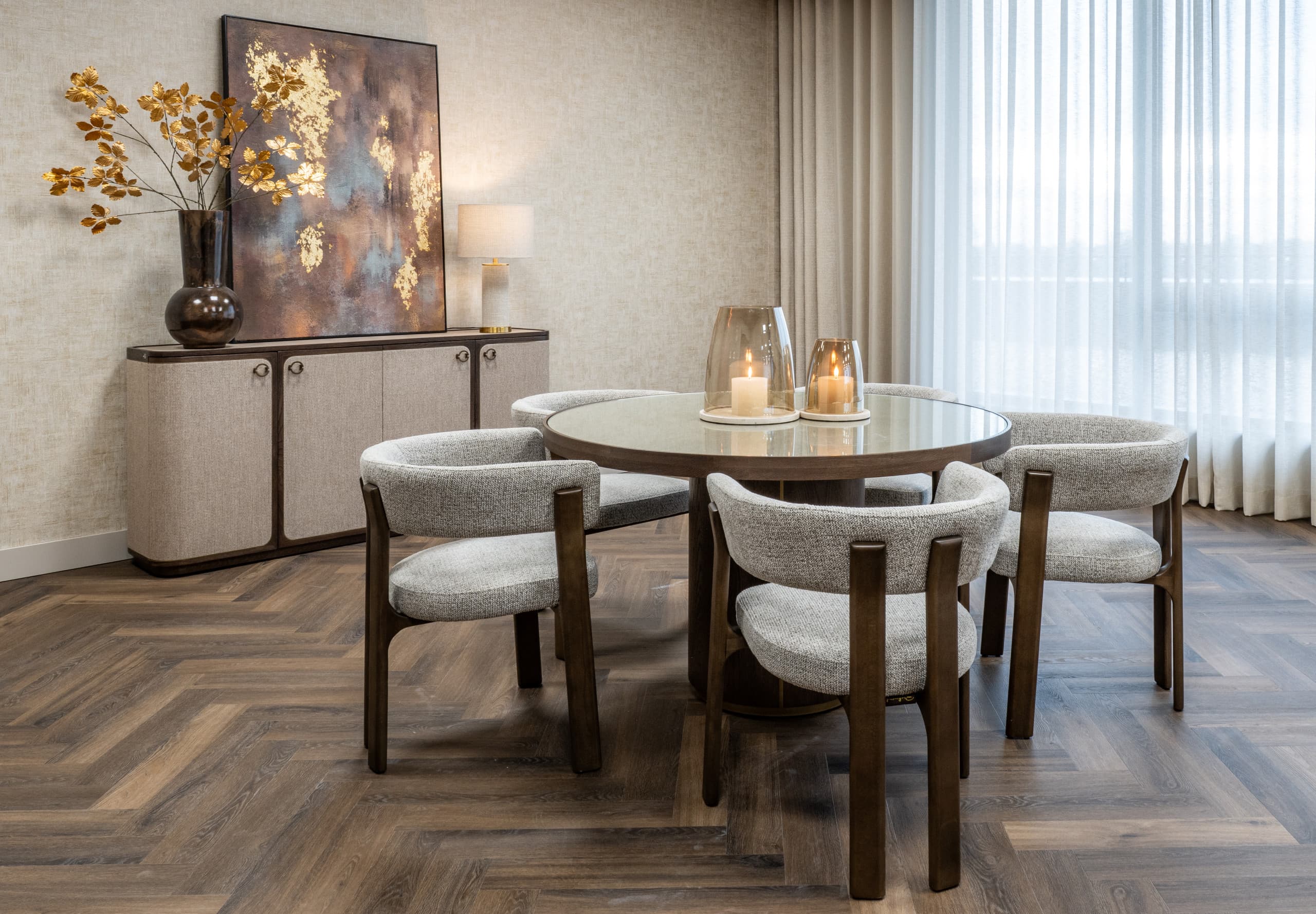 Eetkamer met een goudkleurig abstract schilderij op een dressoir, gouden takken in een vaas en twee windlichten op een ronde tafel