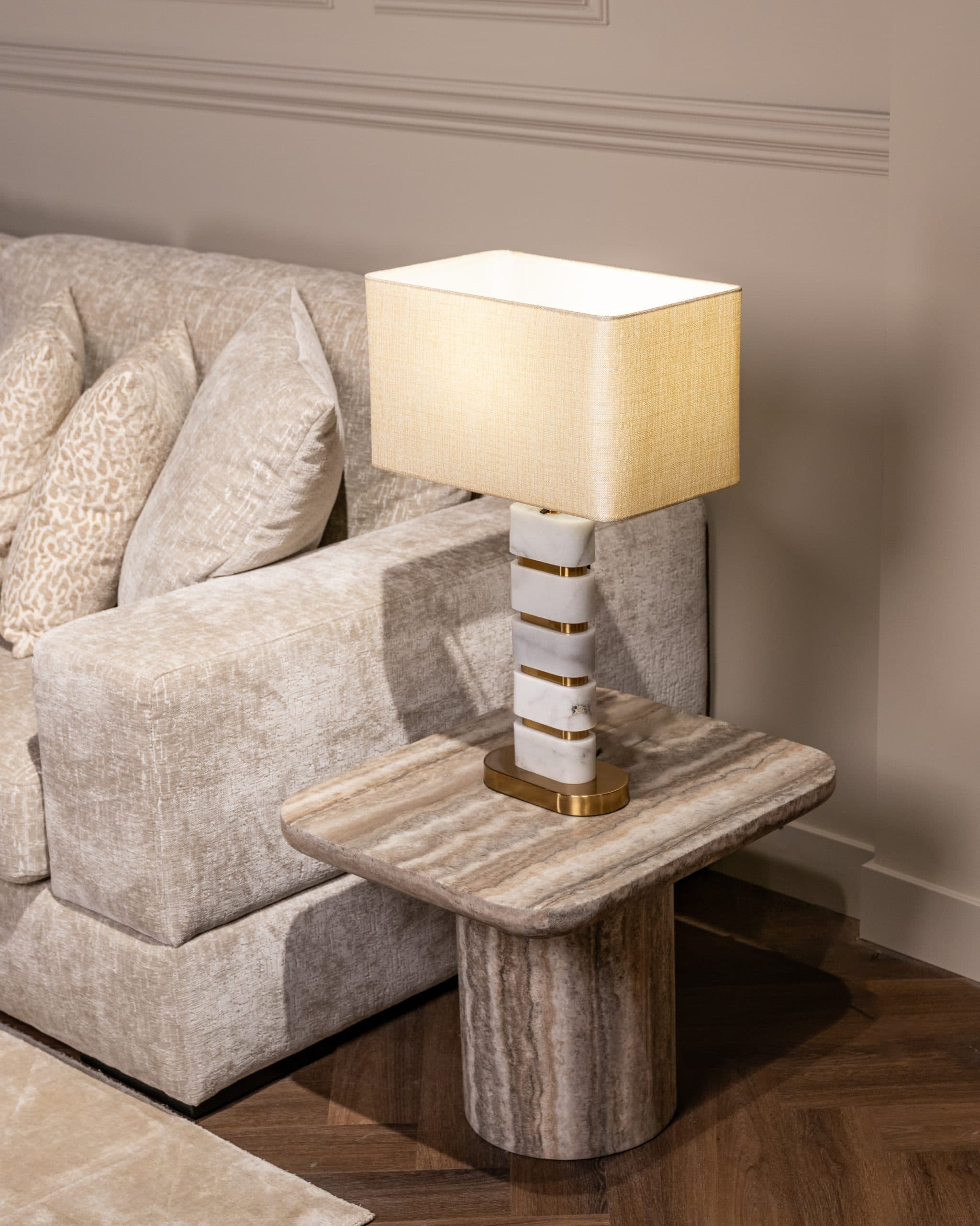 Bijzettafel met travertine blad naast een lichtgrijze bank, met een marmeren tafellamp erop