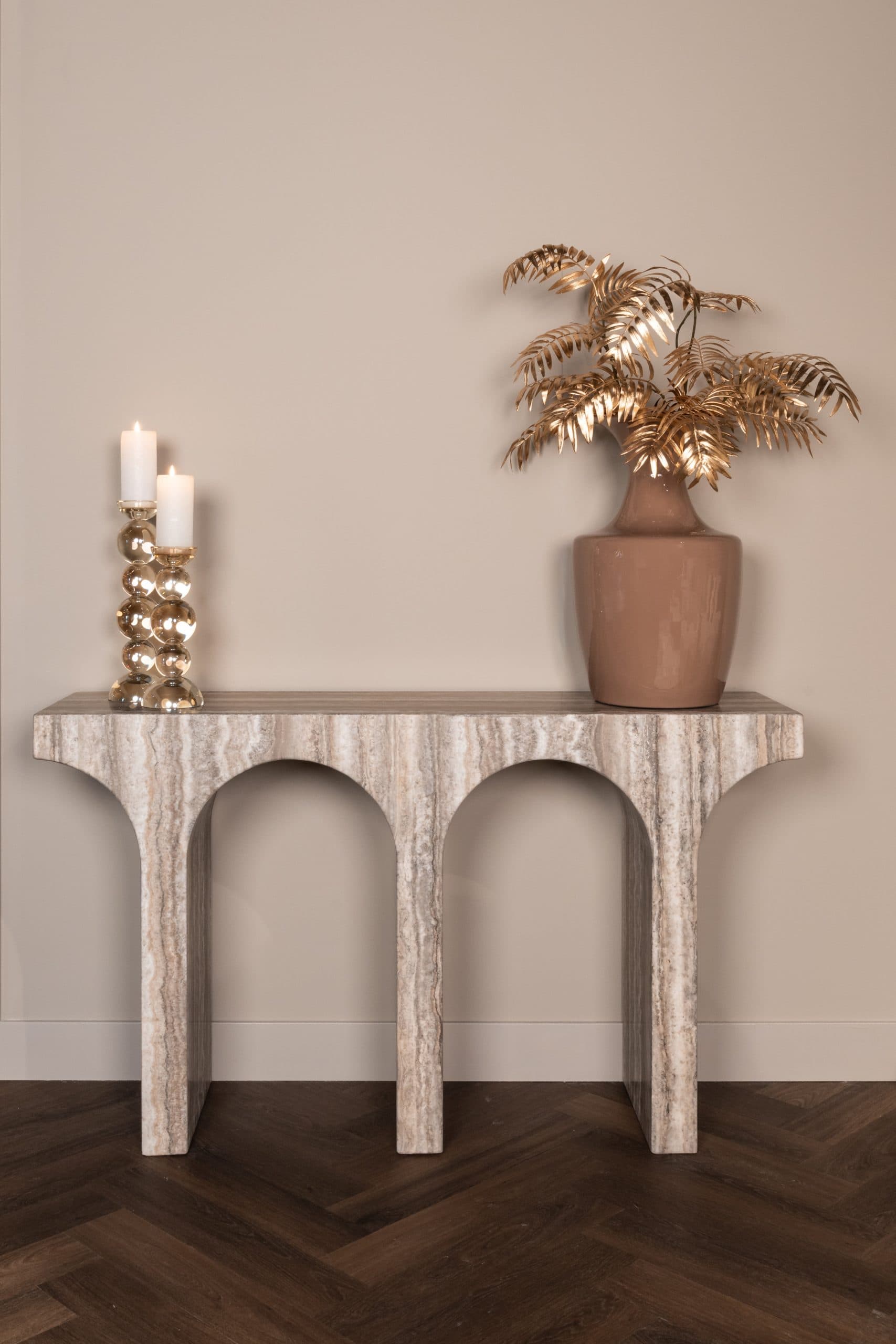 Travertine console met boogpoten tegen een zandkleurige wand, met twee goudkleurige bolkandelaars met witte kaarsen en een terracottakleurige vaas met gouden bladtakken