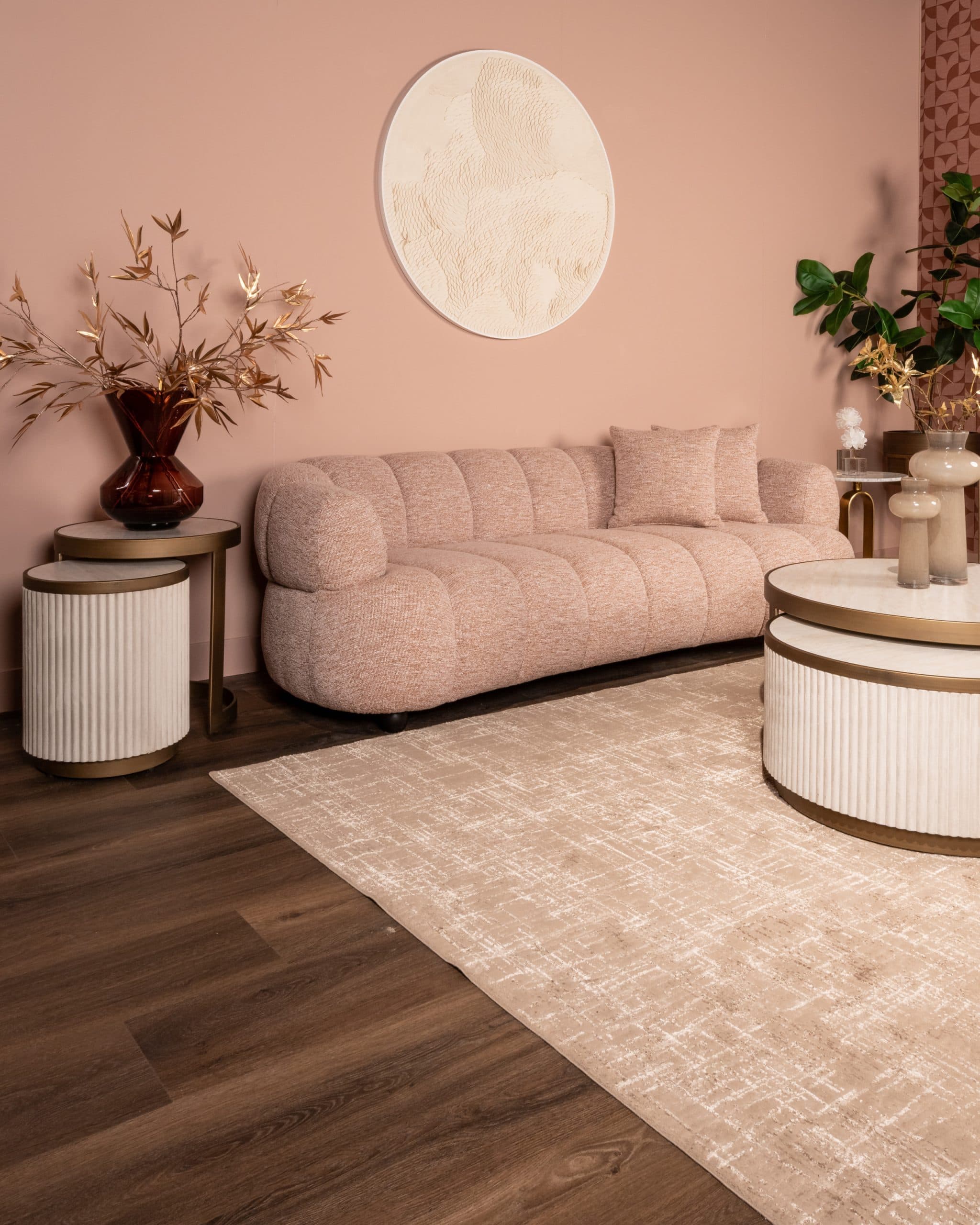 Zacht roze bouclé bank tegen een lichtroze wand, met daarnaast twee ronde bijzettafels met geribbelde voet, een groot beige vloerkleed en geribbelde vazen op de salontafel