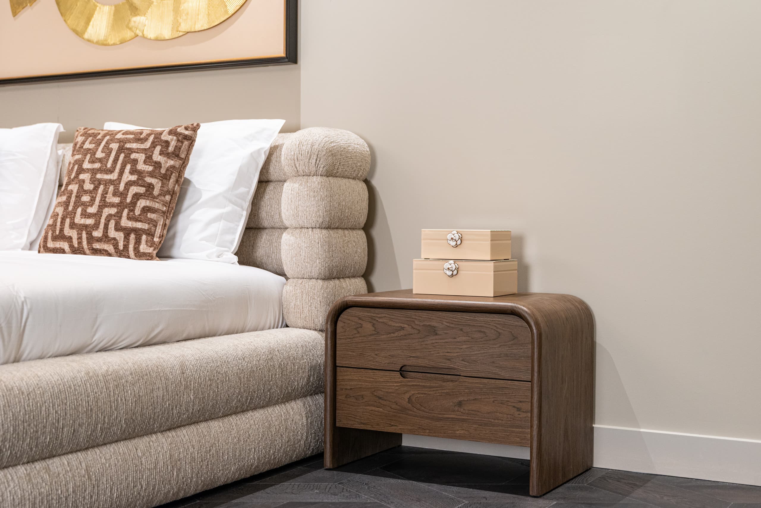 Opgemaakt bed met wit beddengoed en een bruin sierkussen, ernaast een houten nachtkastje met twee beige opbergdozen