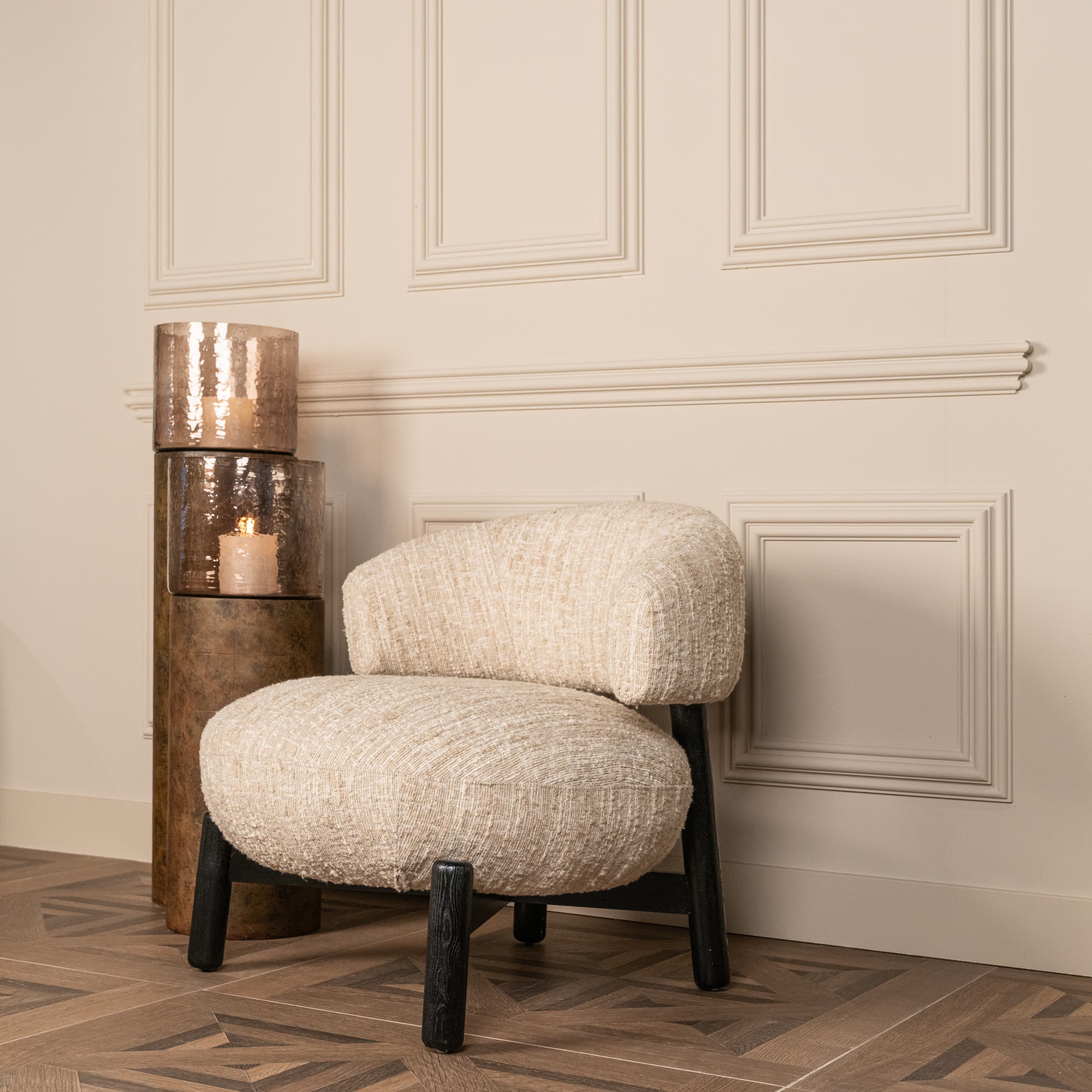 Ronde fauteuil met beige bouclé bekleding naast een pilaar met twee windlichten, tegen een lichte wand met paneellijsten