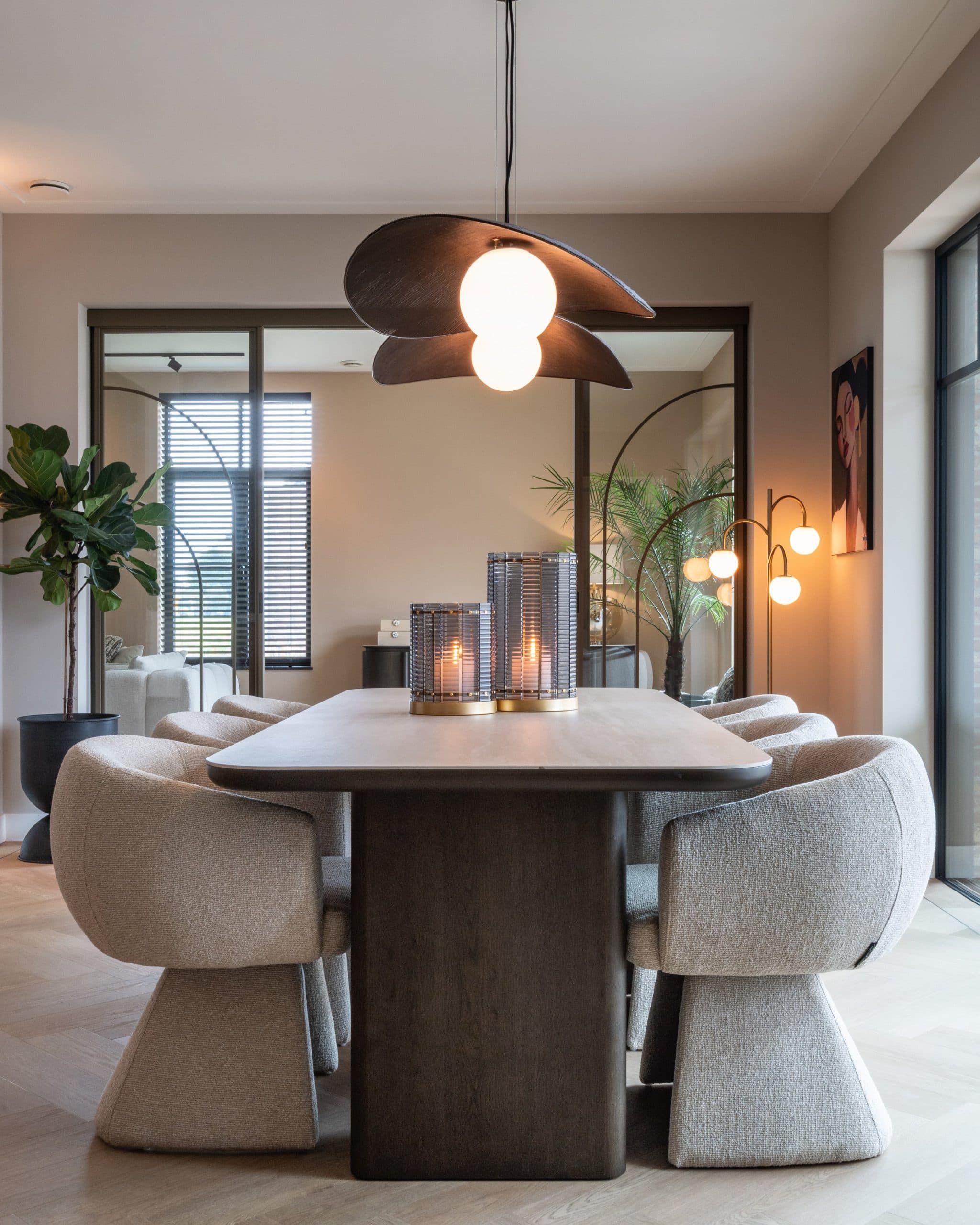 Kale eettafel met donker travertijnlook blad en beige gestoffeerde stoelen, met twee windlichten met brandende kaarsen in het midden en een hanglamp erboven