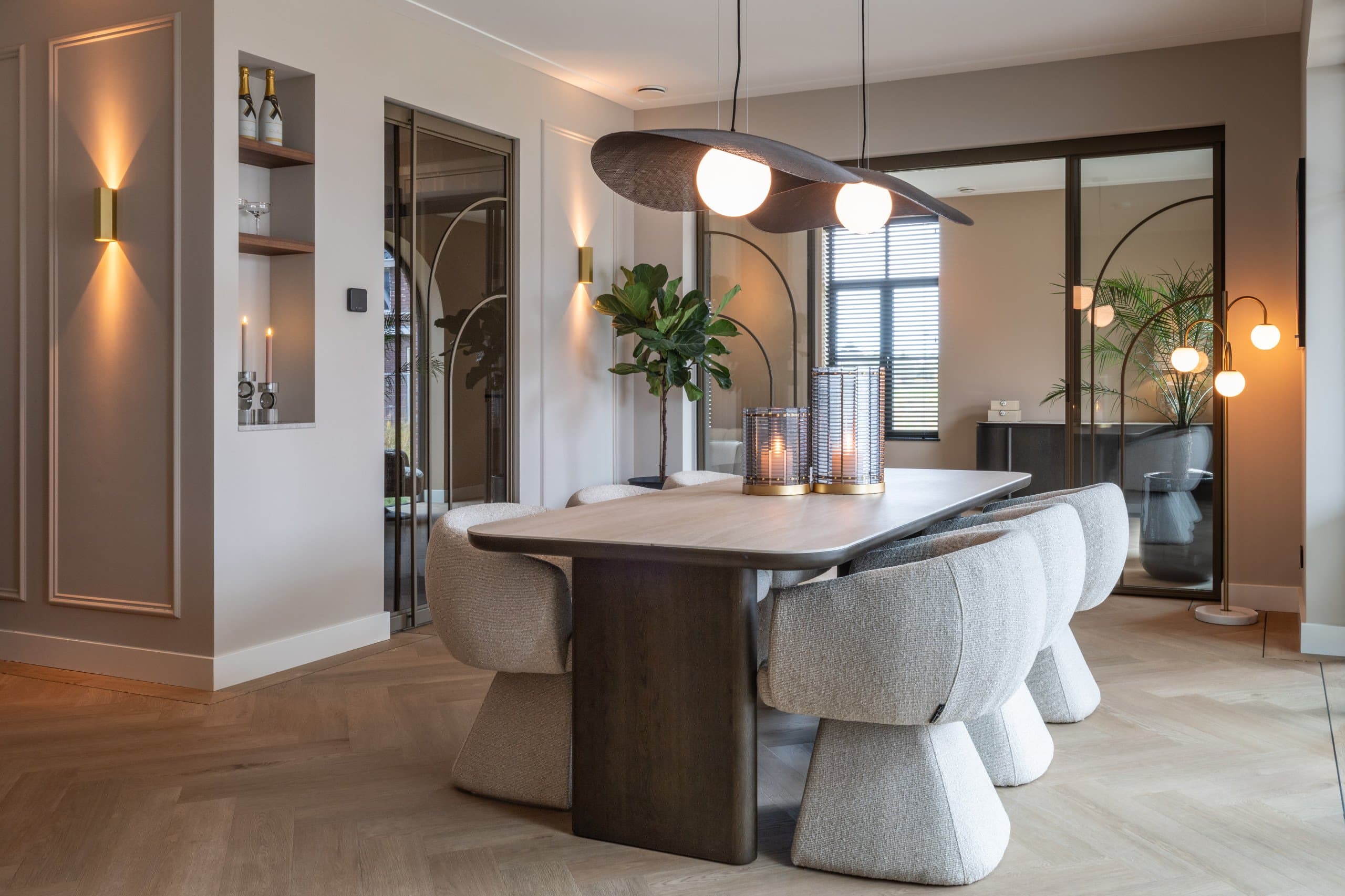 Eetkamer met een houten tafel, bouclé eetkamerstoelen, twee windlichten met brandende kaarsen en wandlampen tegen een taupe wand