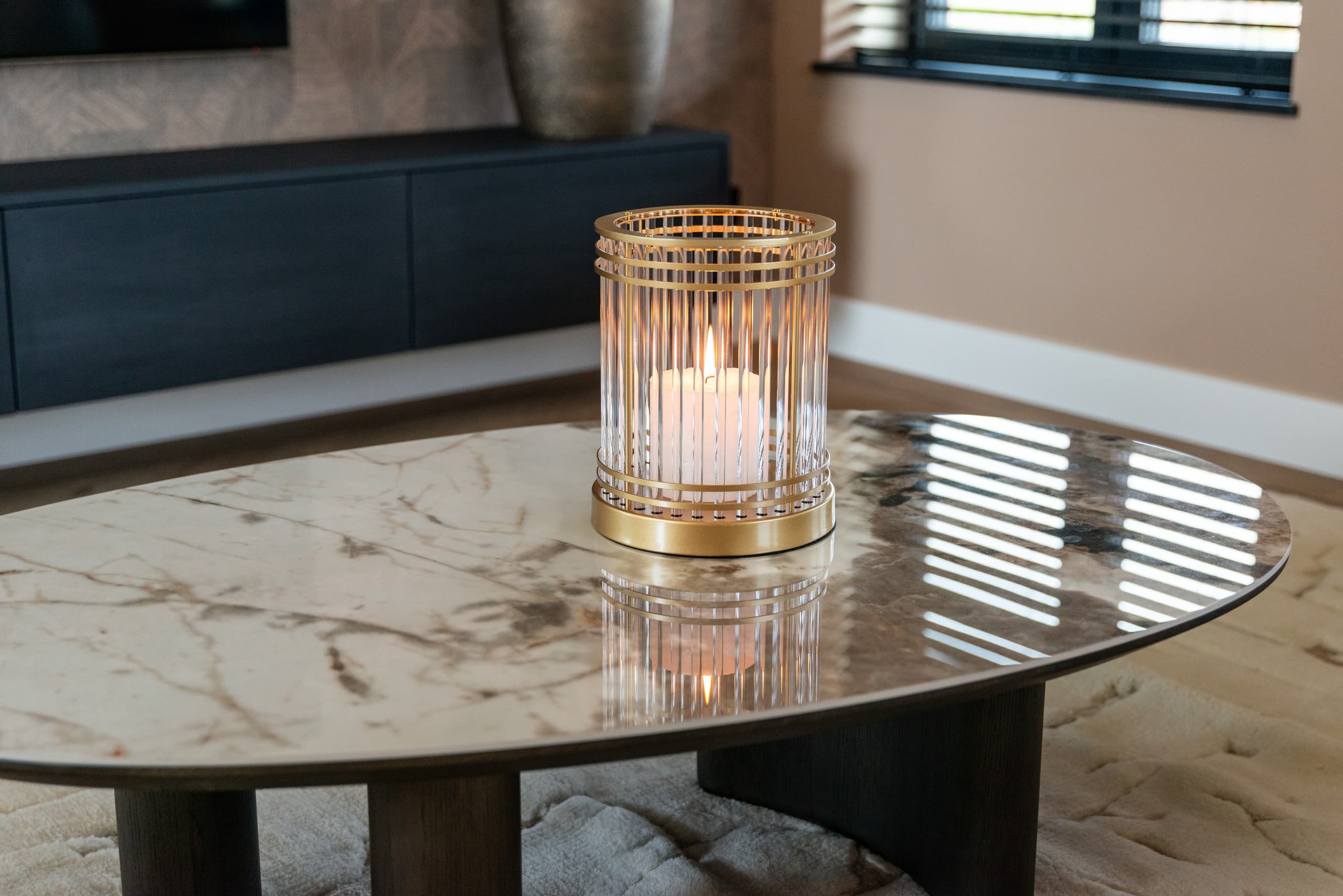 Ovale salontafel met marmeren blad waarop één goudkleurig windlicht met brandende kaars staat, met op de achtergrond een donker dressoir in een lichte kamer