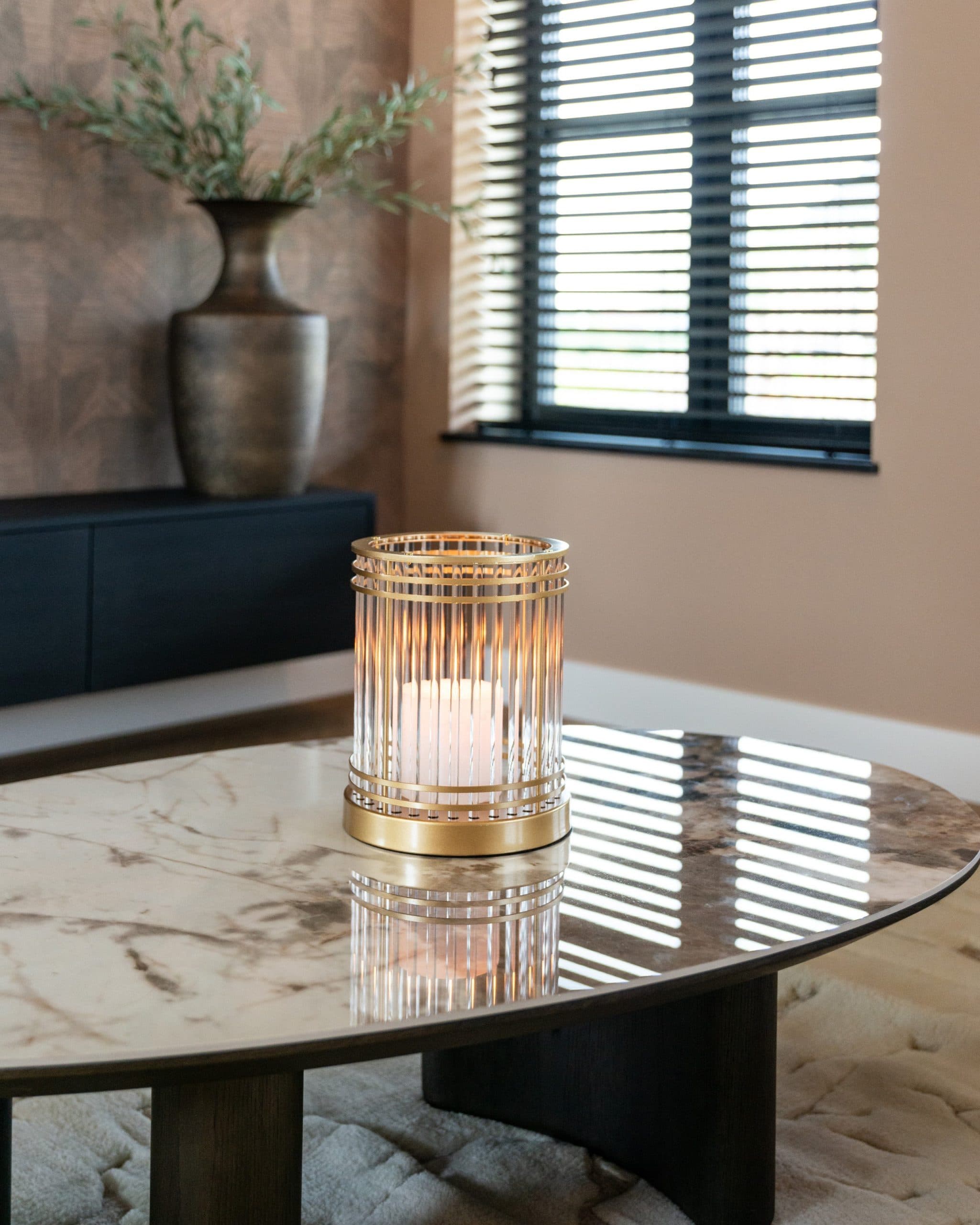 Goudkleurig windlicht met een brandende kaars op een keramisch salontafelblad, met een donker dressoir en een lamellenraam op de achtergrond