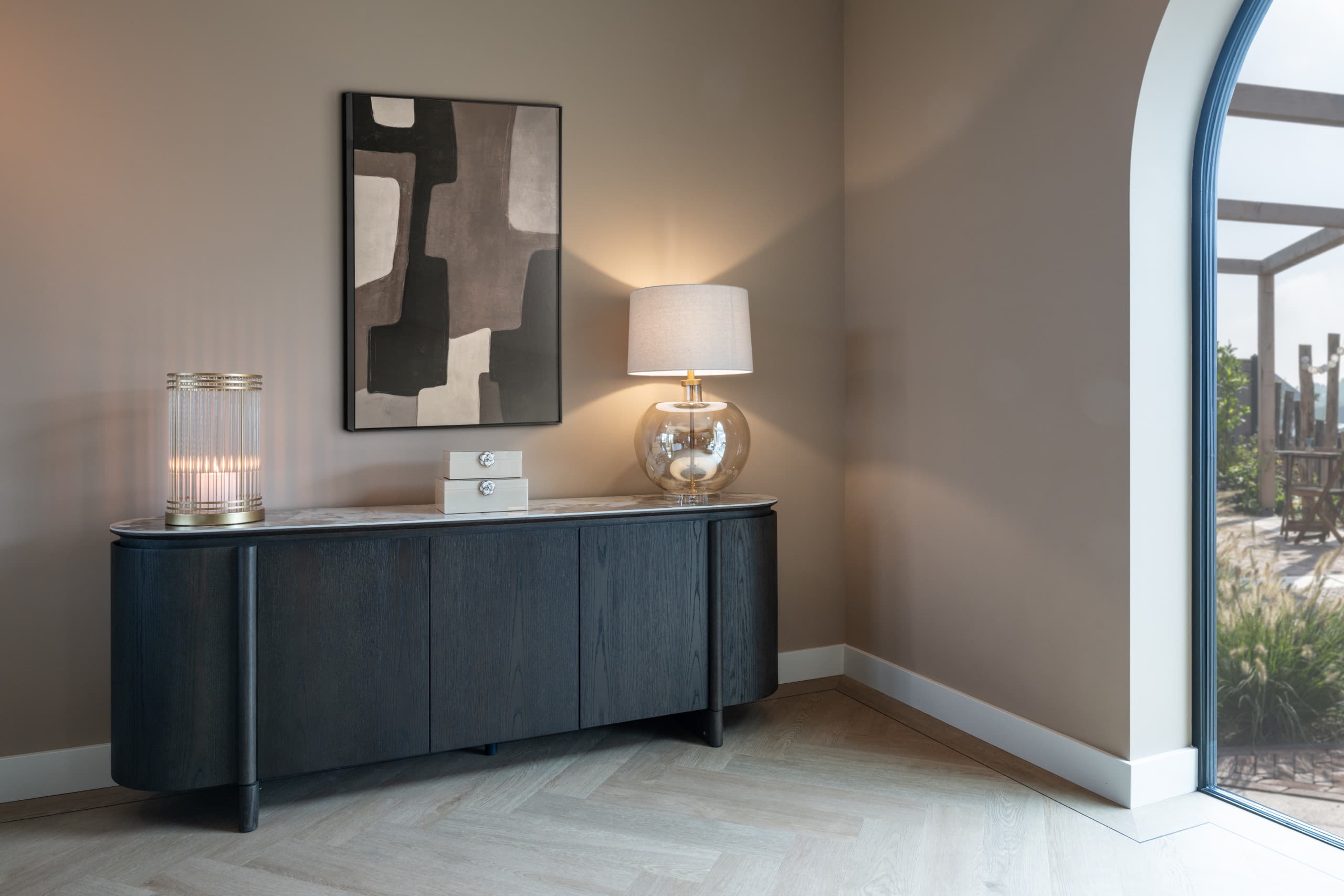 Zwart eiken dressoir met stenen blad, een glazen tafellamp, een windlicht en een abstract schilderij tegen een taupe wand